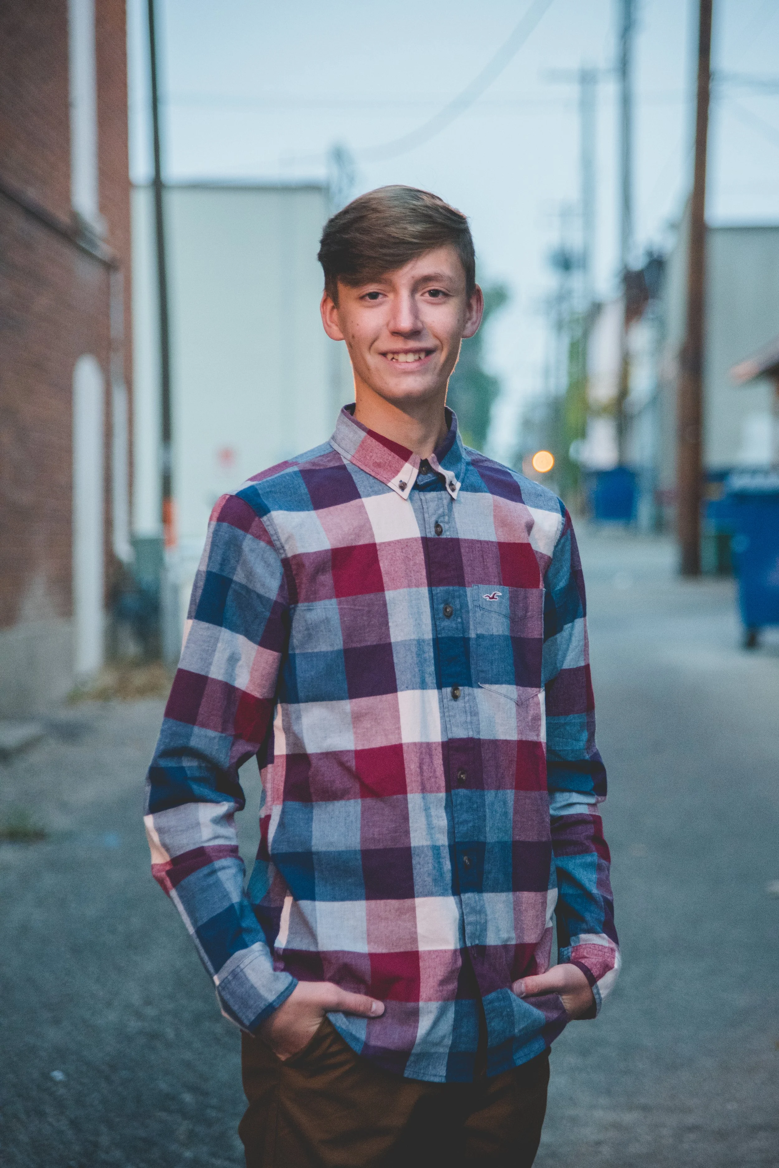 A teenage boy with light skin, dark blonde hair, and brown eyes, wearing a colorful checkered button-up shirt, standing outdoors in an alley at dusk with his hands in his pockets and smiling.