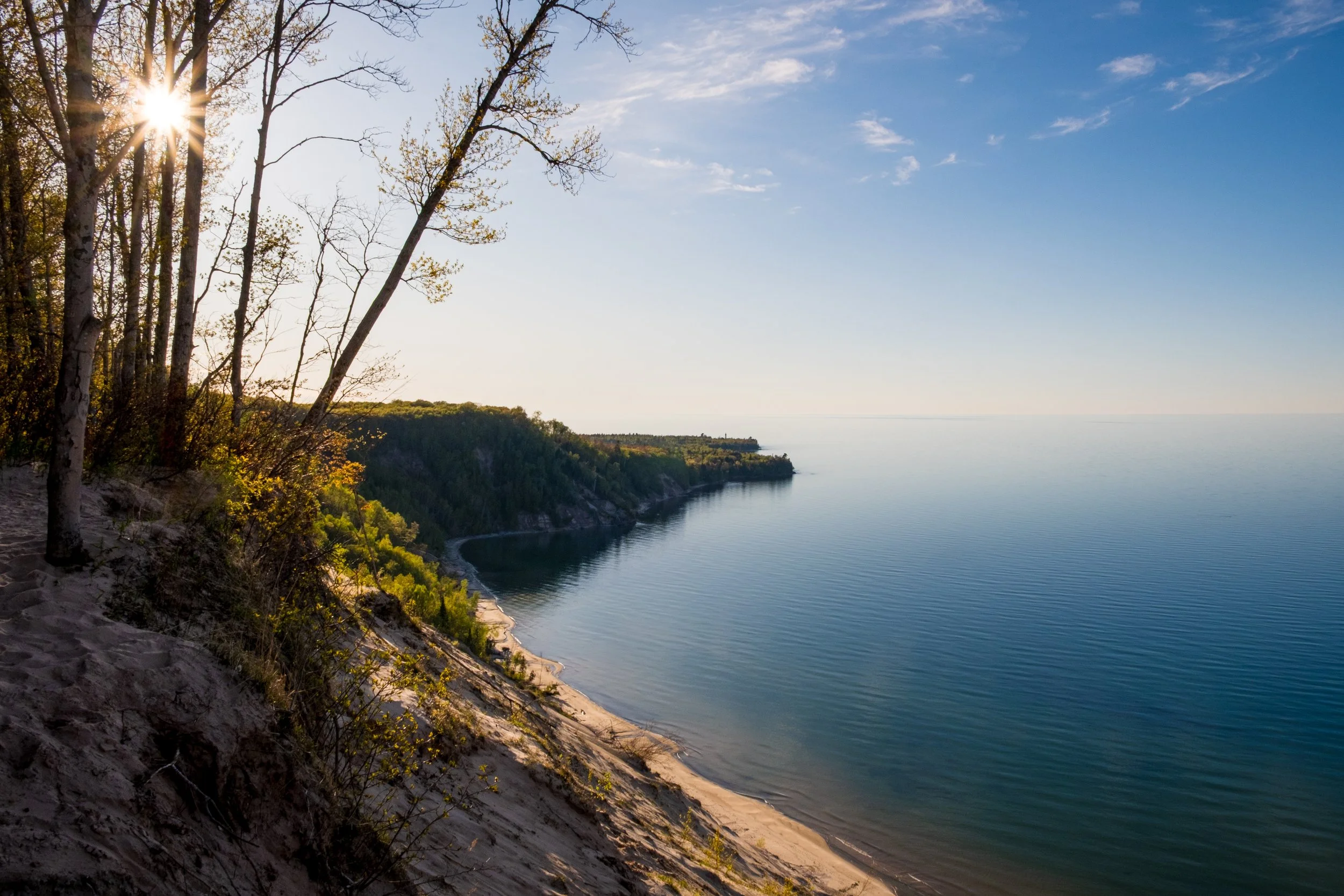 A scenic view of a coastline with a sandy beach, trees, and calm water under a clear blue sky with some clouds. The sun is shining through the trees on the left.