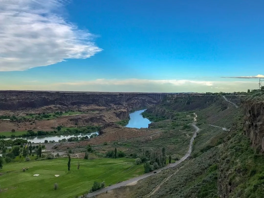 A scenic landscape featuring a winding river flowing through a canyon, with green fields and trees in the foreground and a partly cloudy blue sky overhead.