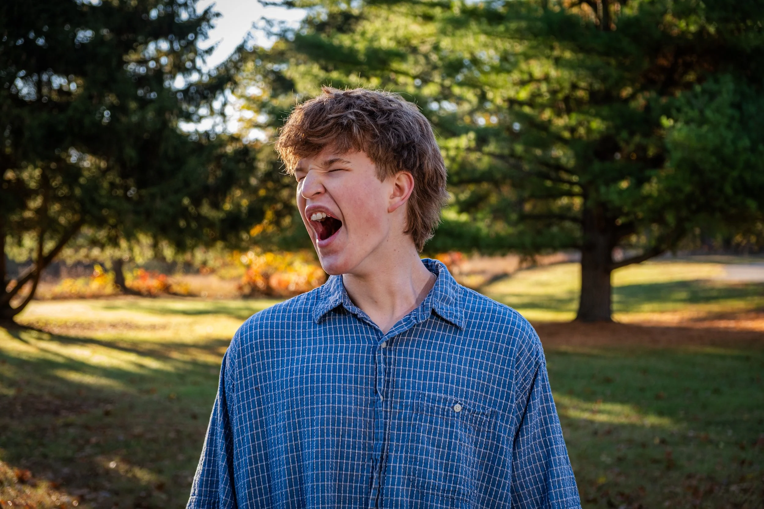 Young man with light brown hair wearing a blue checkered shirt, standing outdoors in a park with green trees and yellow leaves, making a funny face with eyes closed and mouth open.
