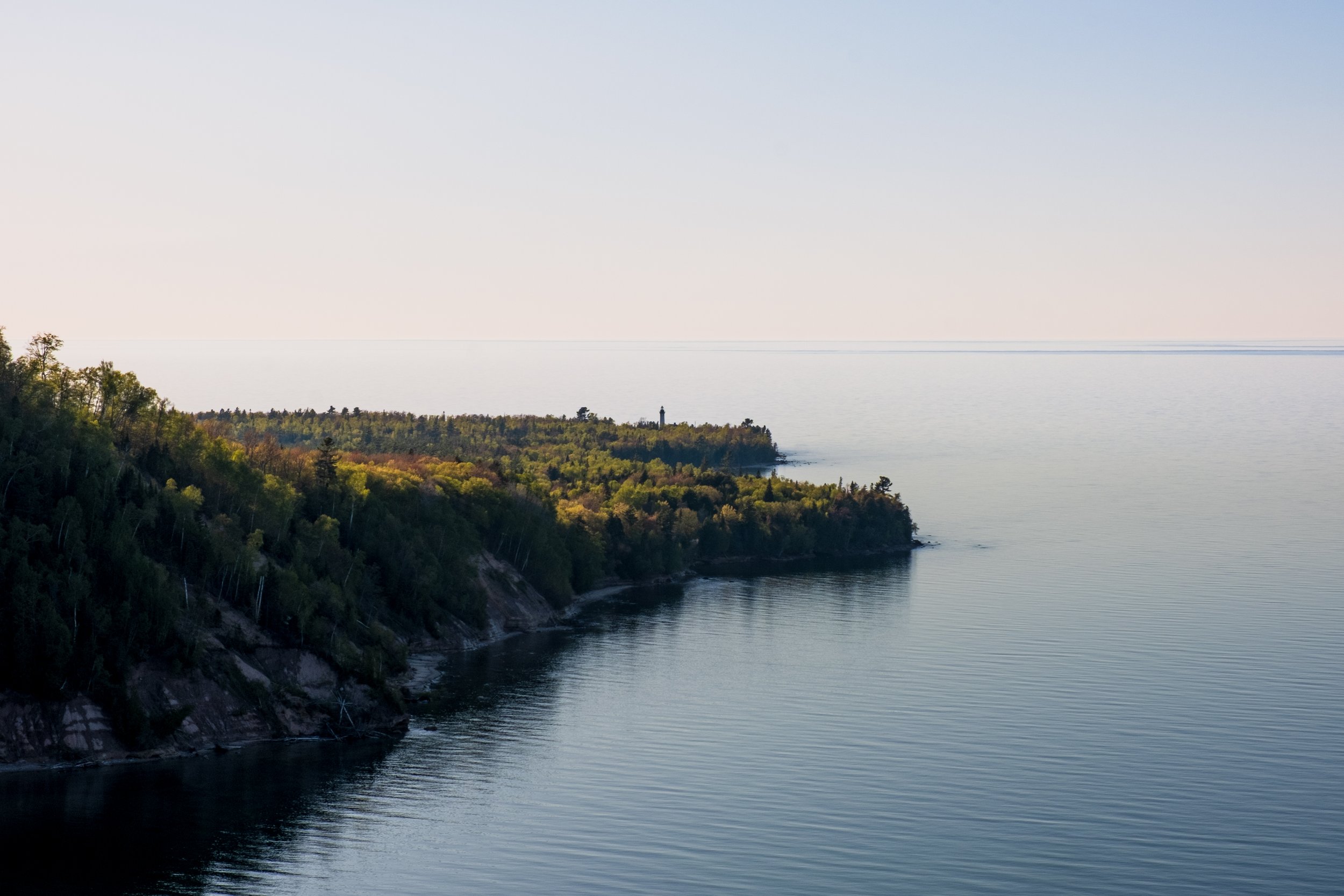 View of a calm body of water with a forested coastline in the foreground and a lighthouse on a small hill on the shoreline.