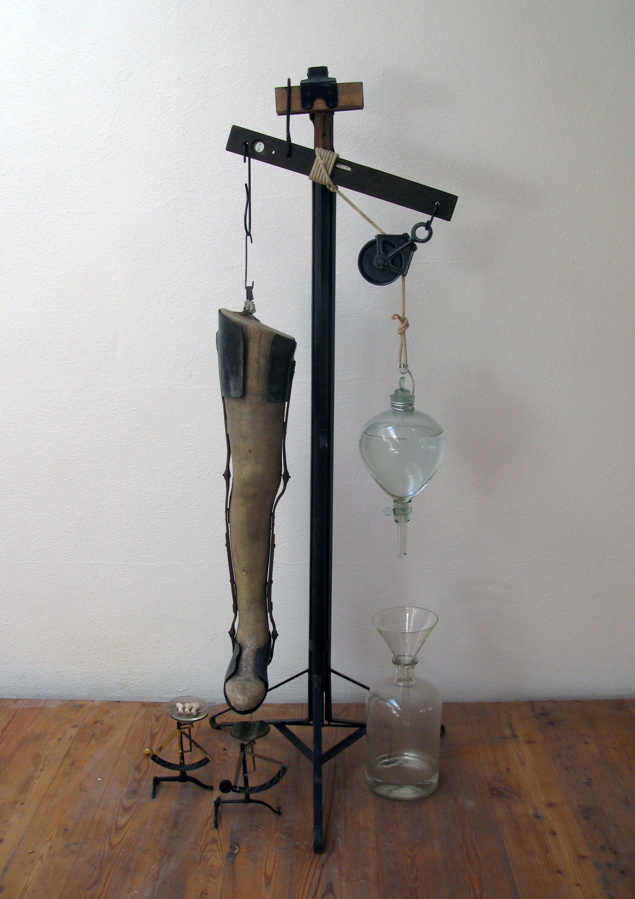 Apparatus for Learning to Step Lightly, Antique leg, tripod, scales, ceramic doll legs, glassware, level, rope, water and hardware. (In this kinetic sculpture, the water drips out of the flask over the course of 4 hours to gently lower the leg onto the scale.) 2014.