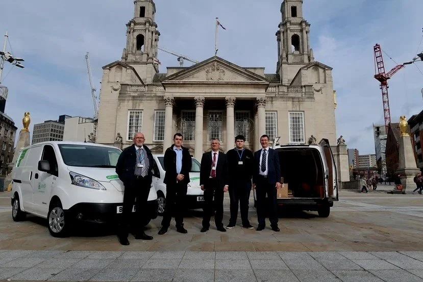 Five men in business attire standing in front of white delivery vans in front of a historic church building, with cranes and city buildings in the background.