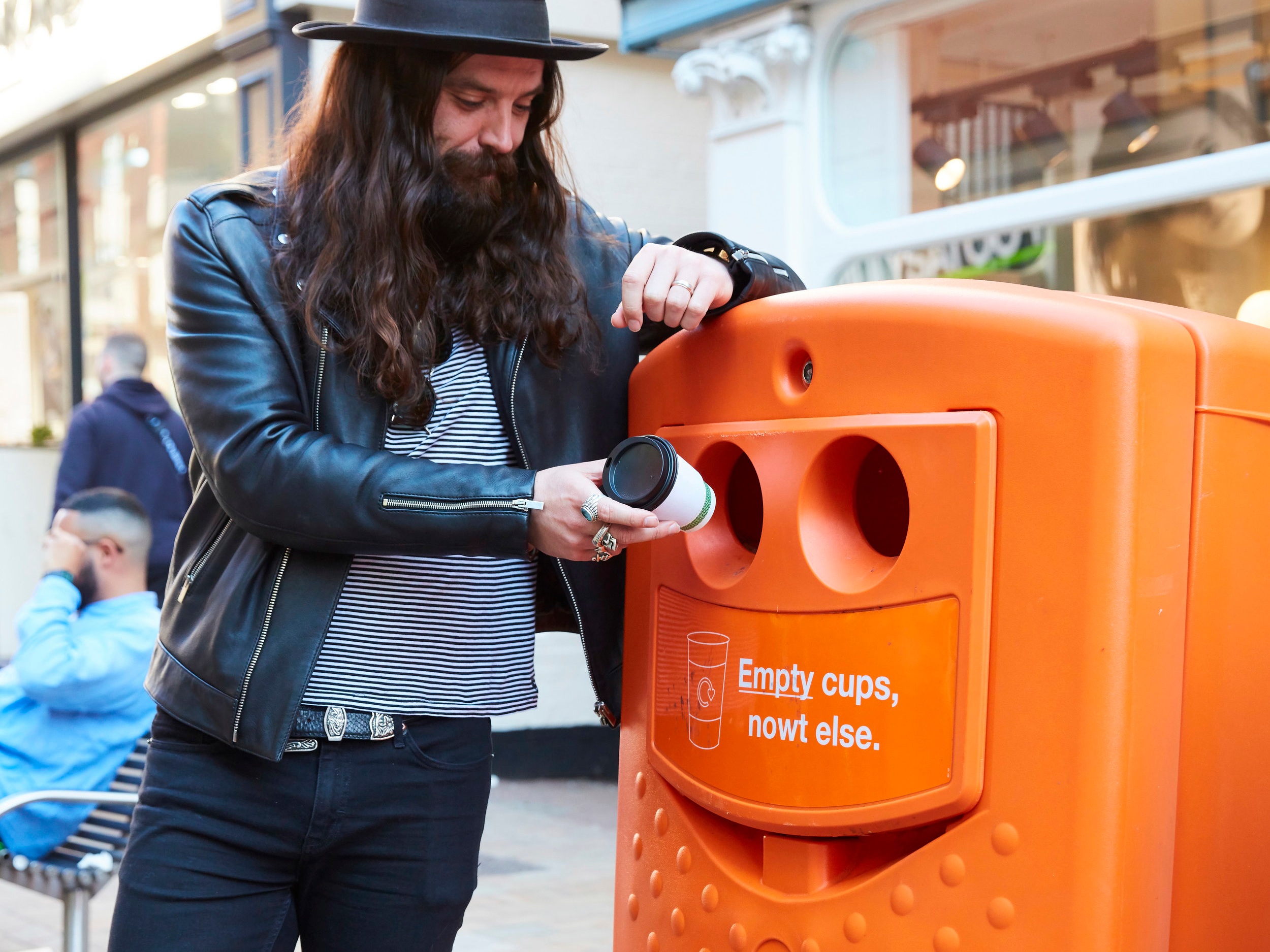 A man with long hair, beard, wearing a black leather jacket and a hat, is discarding a used coffee cup into an orange recycling bin labeled for empty cups.