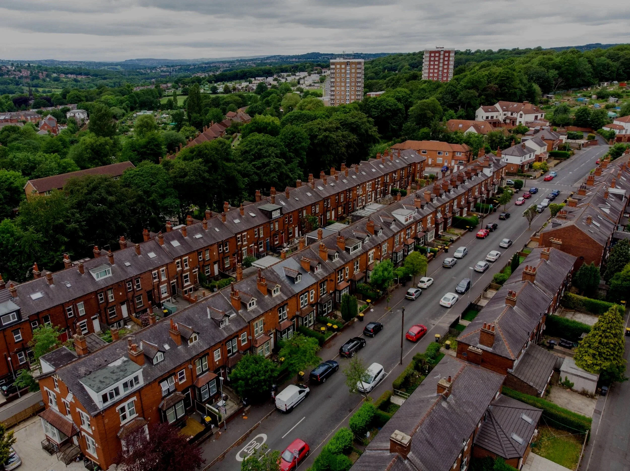 Aerial view of a residential neighborhood with rows of brick houses, parked cars, and green trees, with a hilly landscape in the background under a cloudy sky.
