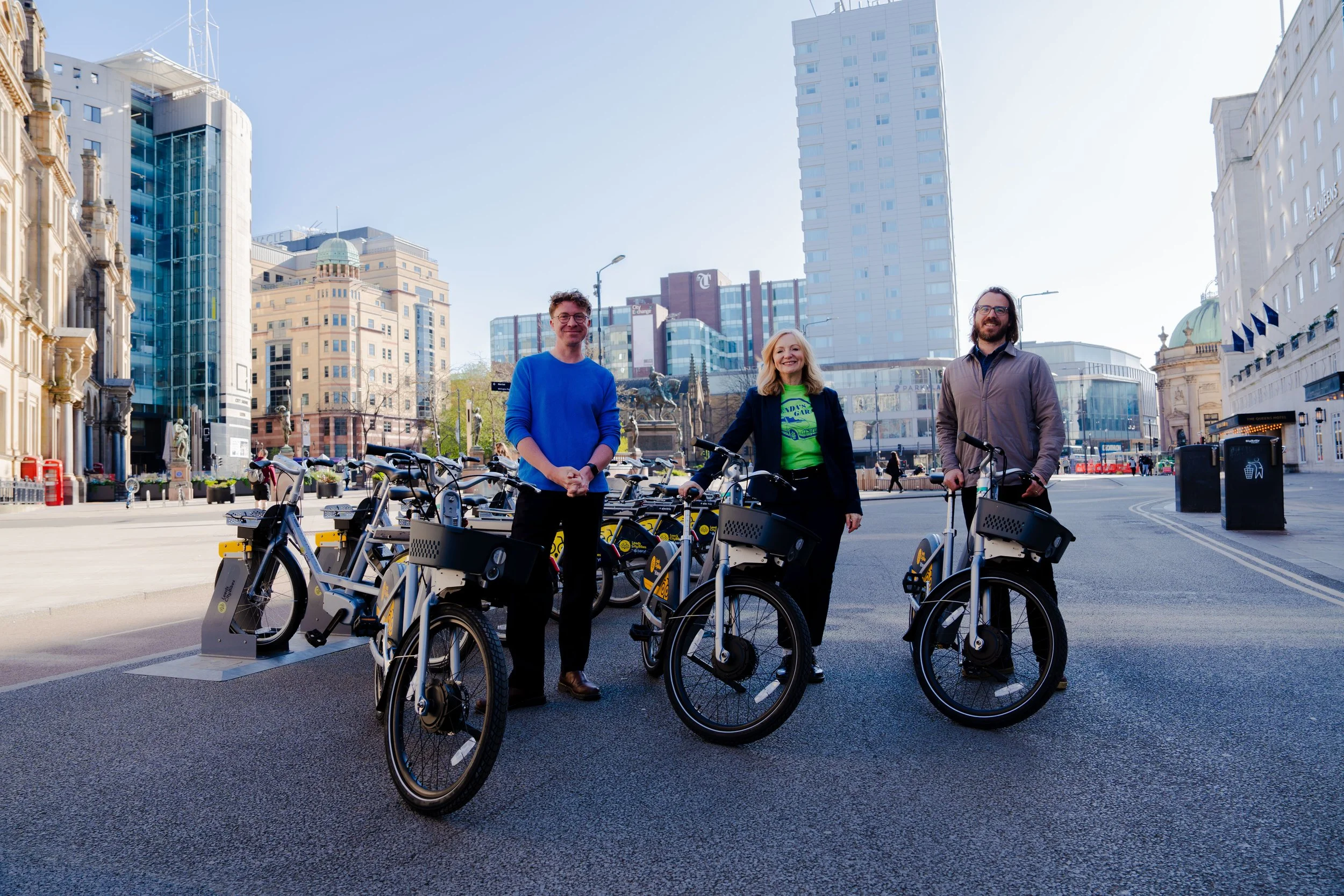 Three people standing with bicycles and e-scooters on a city street during daytime, with tall buildings and a clear sky in the background.