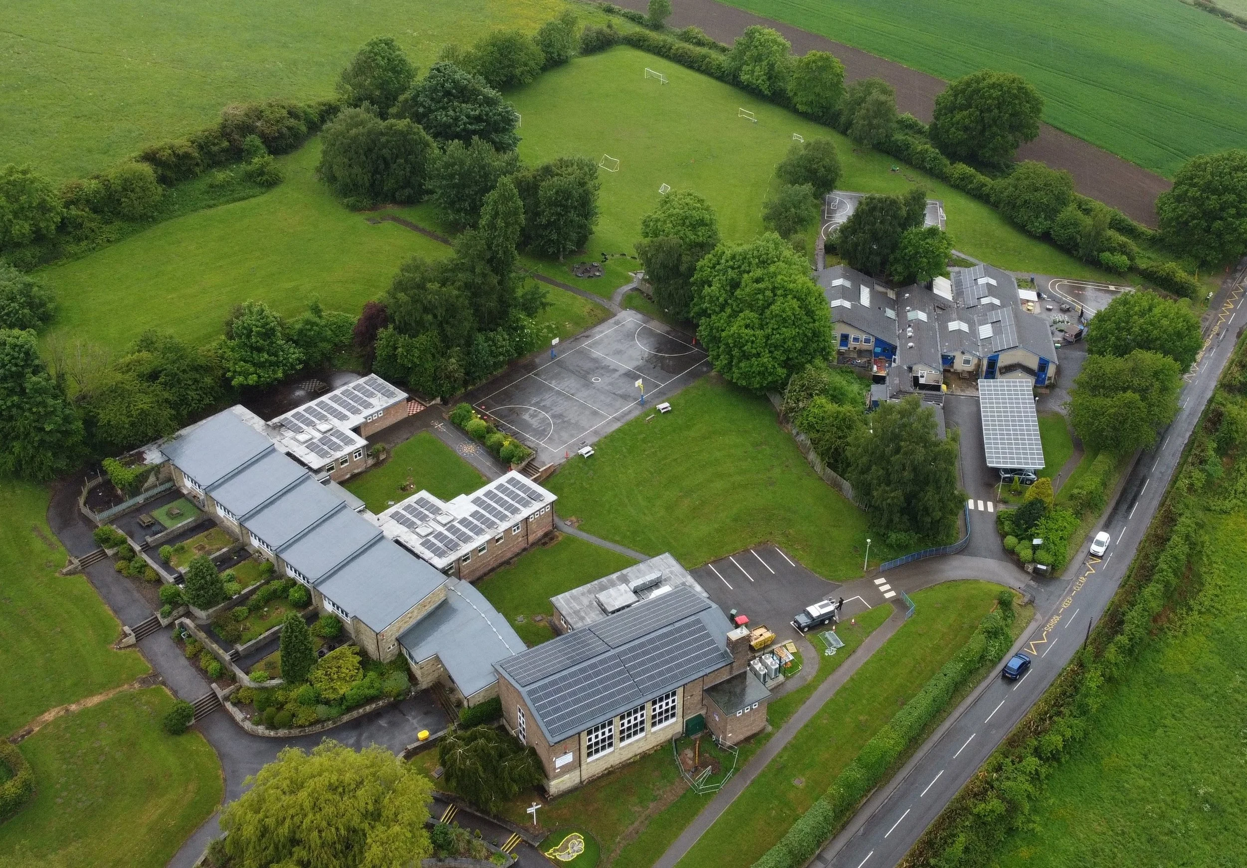 An aerial view of a school campus surrounded by greenery, with shaded trees, playgrounds, sports courts, and parking areas.
