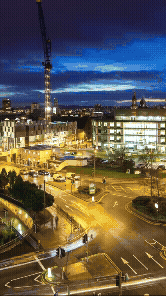 Nighttime cityscape with construction crane, illuminated buildings, and busy streets with traffic.