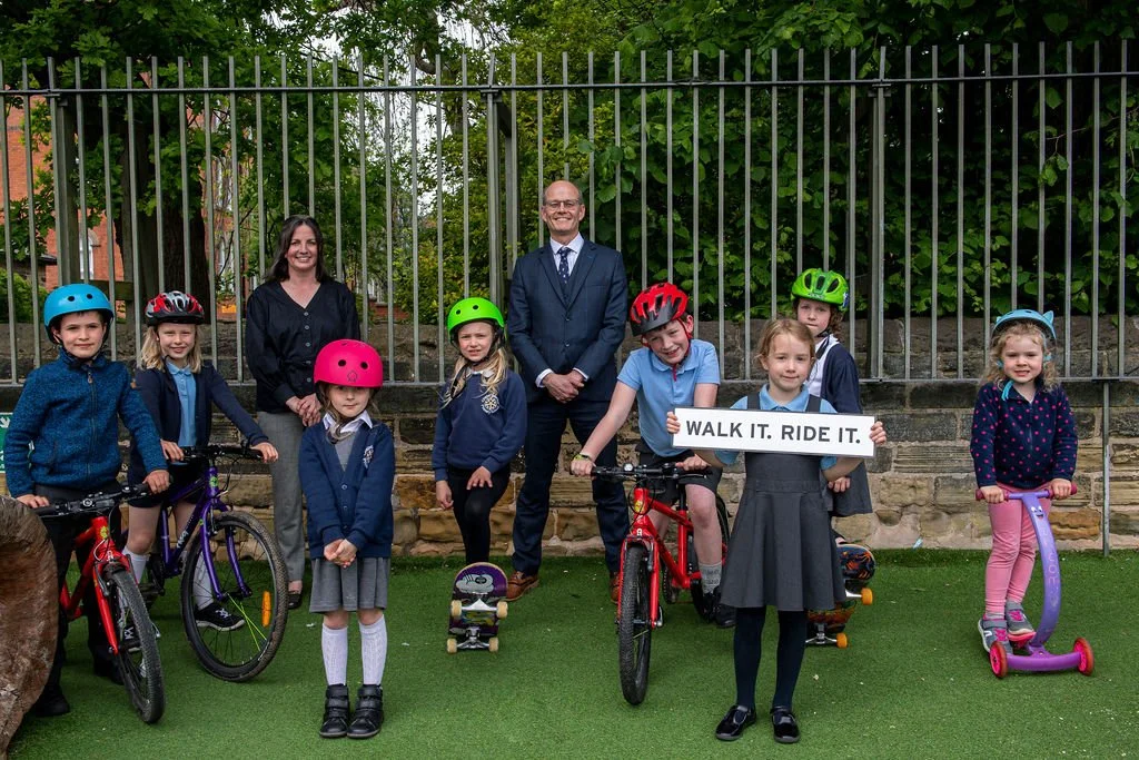 Group of children wearing helmets with bicycles and scooters outdoors, two adults standing behind them, one woman and one man in a suit, holding a sign that reads 'WALK IT. RIDE IT.' on a grassy area with a stone wall and metal fence in the background.