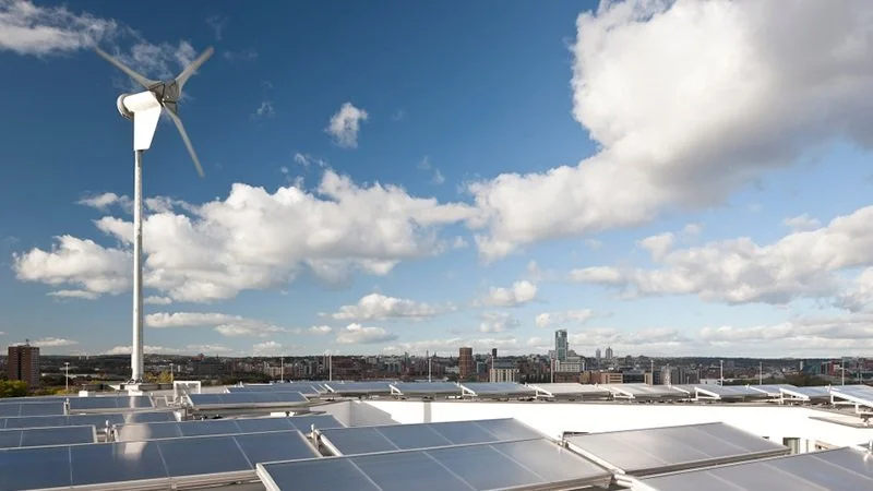 Rooftop with solar panels and a wind turbine in front of a city skyline under a partly cloudy sky.