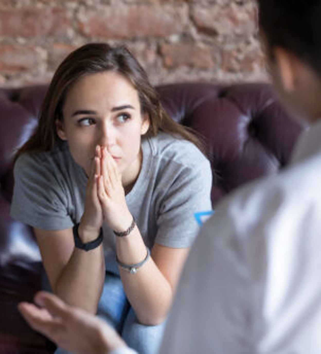A young woman with brown hair, wearing a gray t-shirt, is sitting on a purple leather couch with her hands pressed together in front of her lips, looking attentively at a person who is partially visible and speaking to her, against a brick wall background.