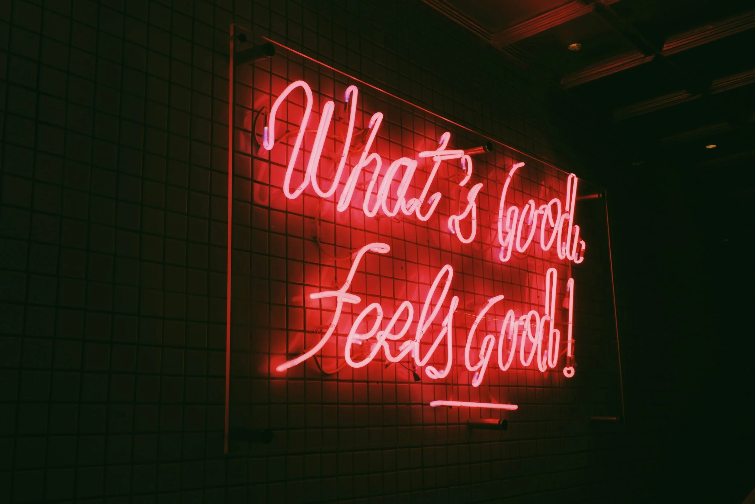 Pink neon sign with a red glow on a tile wall at night that says "What's Good, Feels Good!"