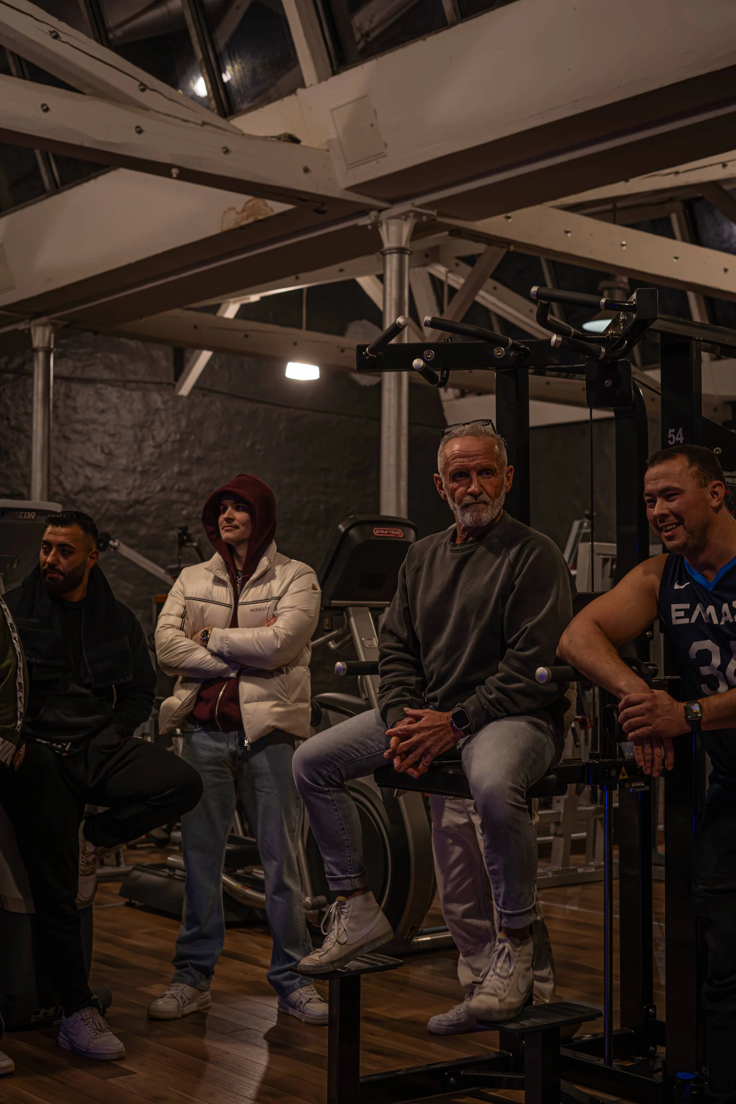 Vier Menschen im Fitnessstudio, einige sitzen auf Sportgeräten, andere stehen, dunkle Beleuchtung, moderne Fitnessgeräte im Hintergrund.