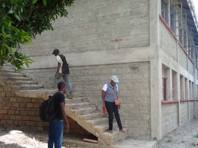 Executive Director, Loyce, is walking down the concrete stairs of the Mwanzo two-story school building while a workman is walking up the stairs.