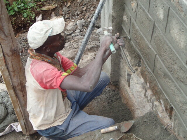 A skilled laborer is installing what looks like a plumbing line from the exterior of the school near the foundation.