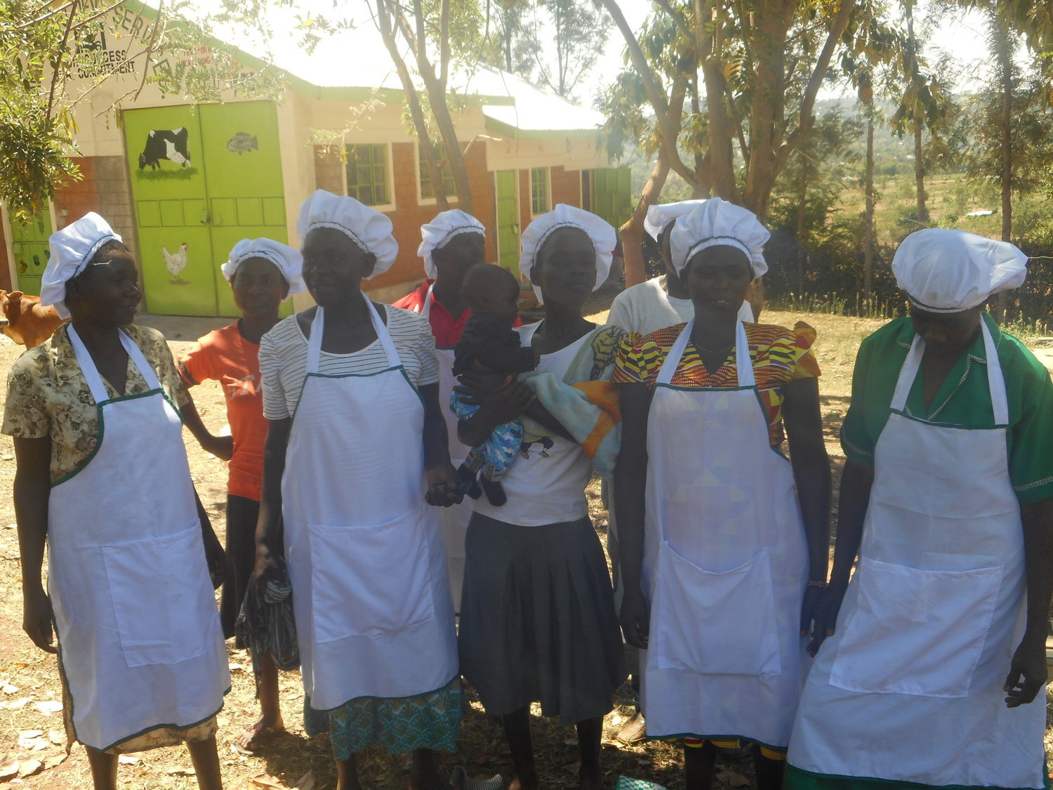 Eight village women who are in the microfinance Woman’s group have gathered with their chef’s hats and aprons in a yard with trees and colorful buildings. One of them is holding a baby.