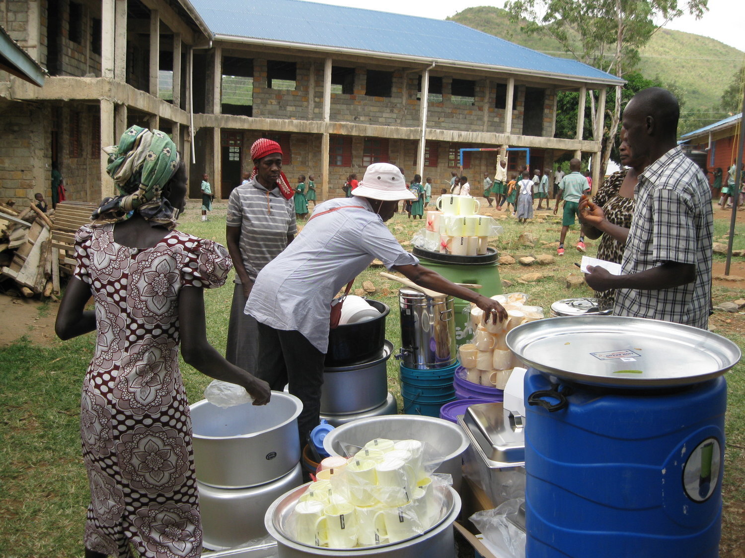 There are many children in the school yard background and five  village persons who are catering an event.  They are seen setting up cups and beverages to be served.