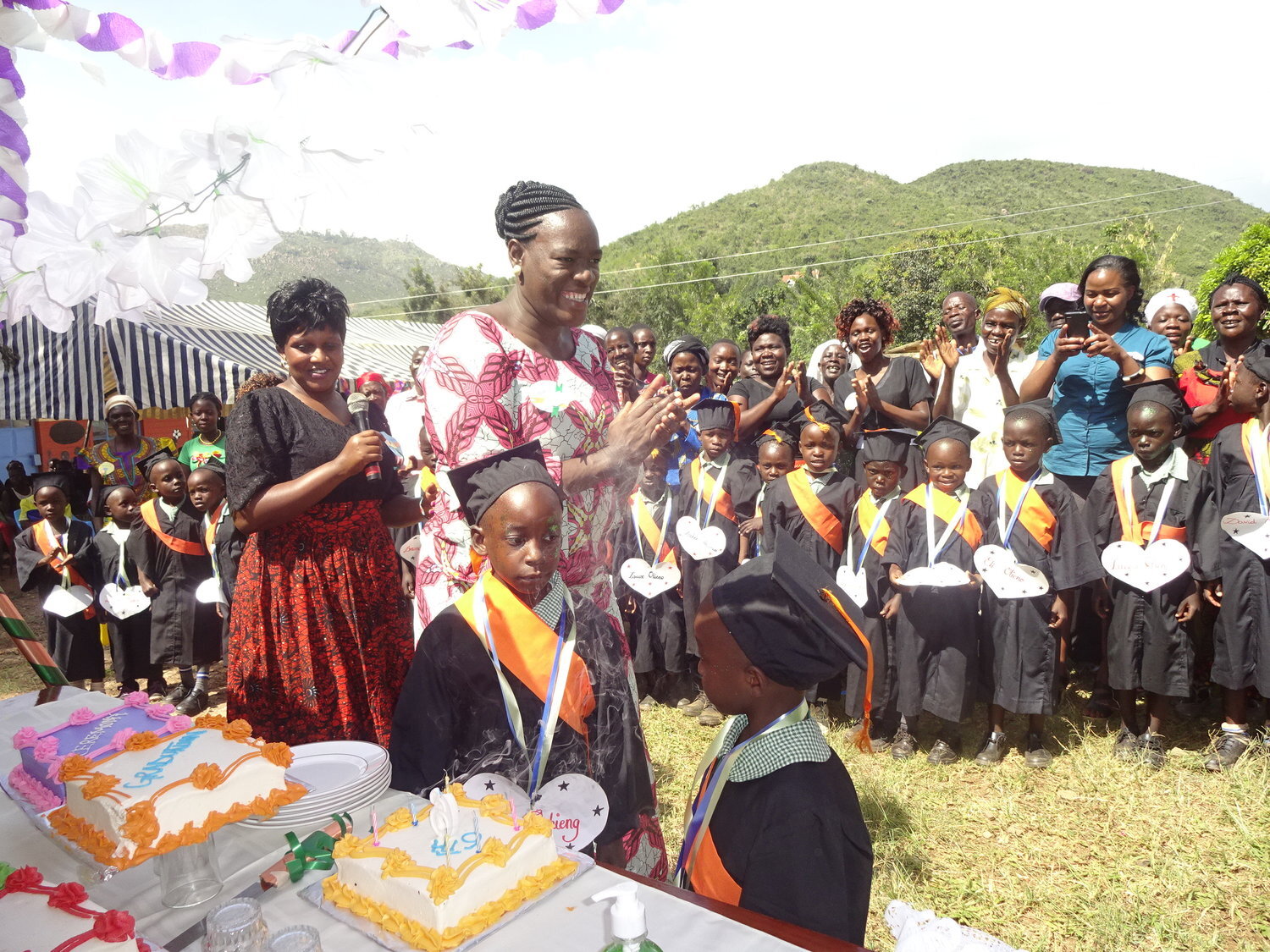 Two young students in graduation regalia stand next to a table with decorated cakes on it while two women clap and an audience including other child graduates looks on