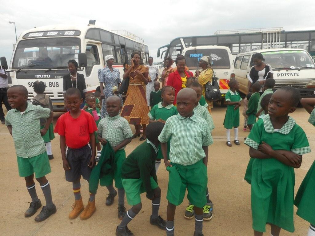 A group of schoolchildren and adults stand in front of three white buses