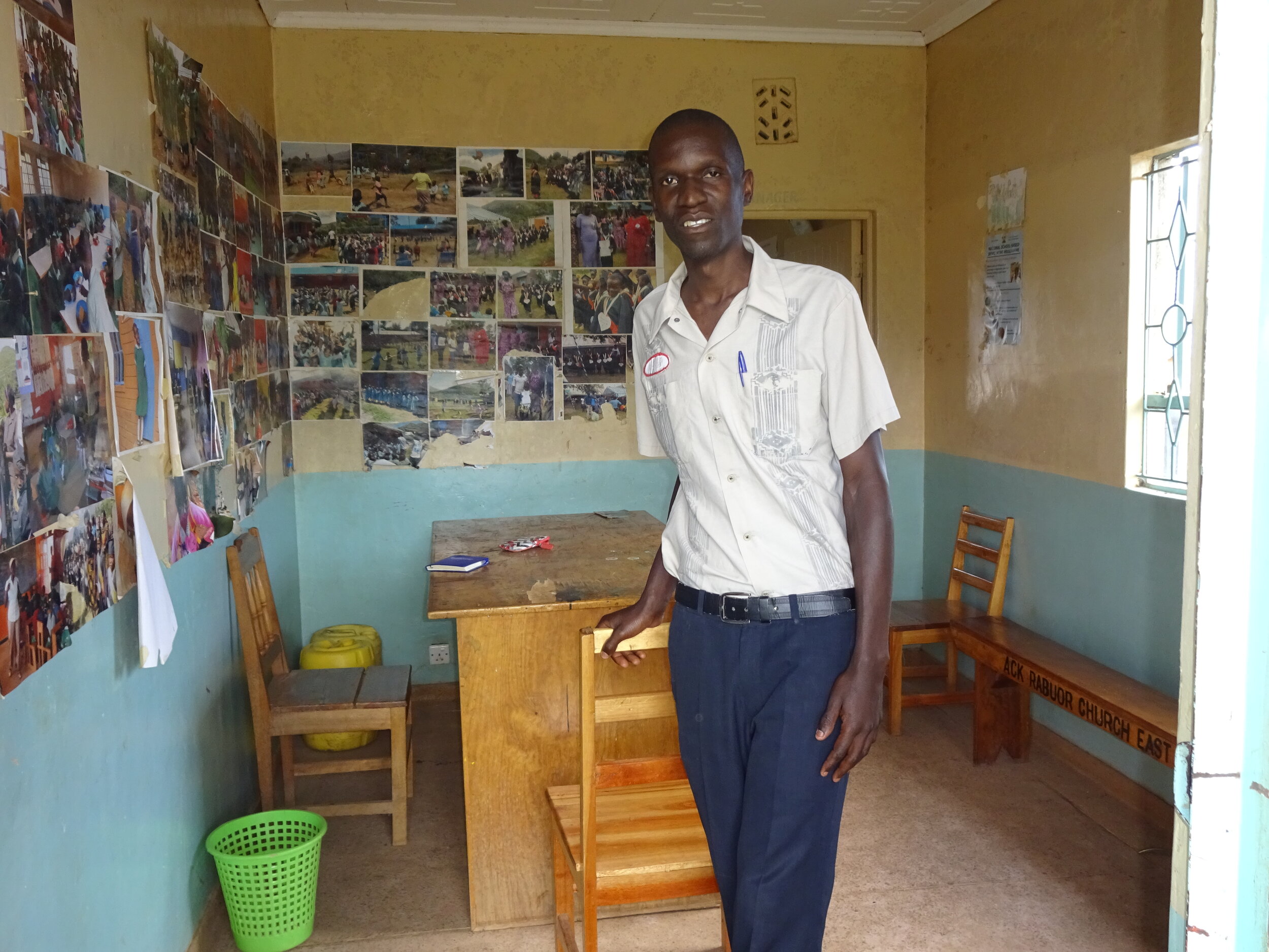 Kevin, one of the Mwanzo teachers, is standing in a small room filled with pictures on the walls and with a desk and two chairs. Sunlight is streaming in through a window.