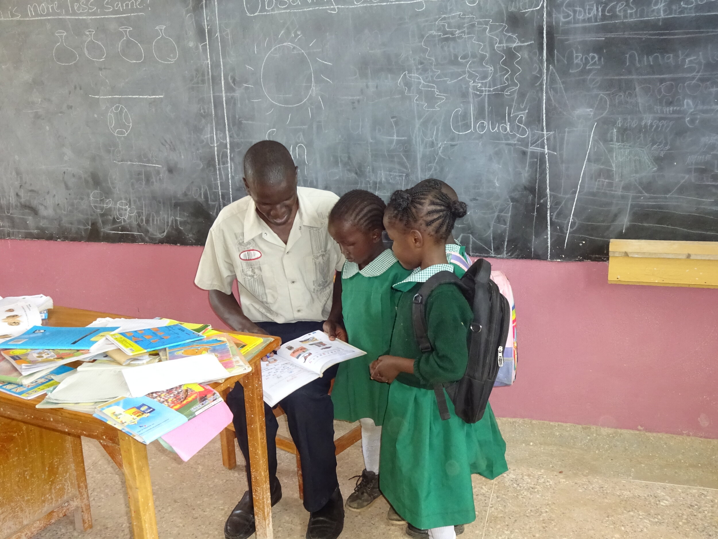 Kevin, a Mwanzo teacher, is  seated in front of a chalk board at a desk filled with many books. He is showing a lesson to two school girls.