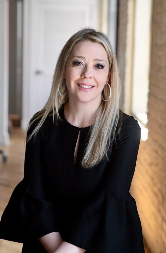 A woman with long blonde hair wearing a black top with a keyhole neckline, smiling, in a room with a brick wall and wooden floors.