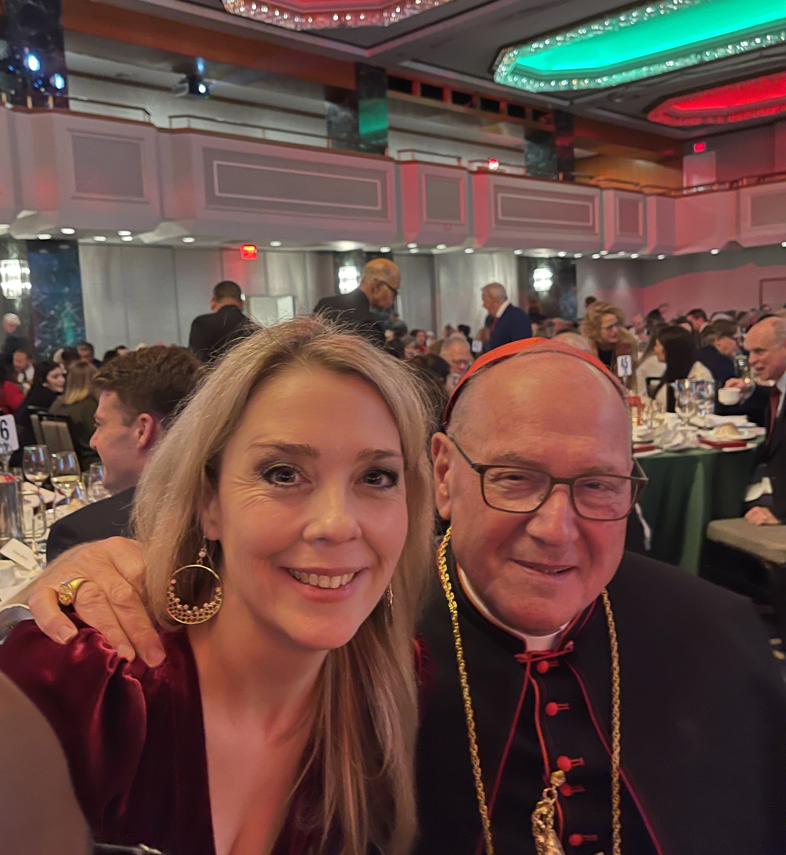 Carly York posing with Cardinal Timothy Dolan in a banquet hall, which is lively with people dining and colorful ceiling lights.