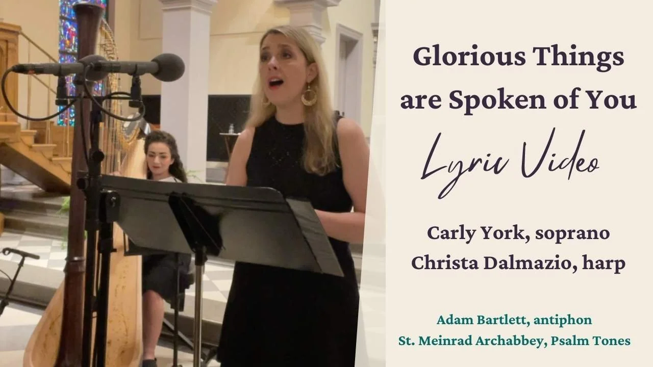 A woman singing into a microphone during a performance at a church. A young girl plays the harp in the background. There is a poster next to the performers with text about a video titled 'Glorious Things are Spoken of You' featuring Carly York and Christa Dalmazio, with additional details about musical pieces and performers.