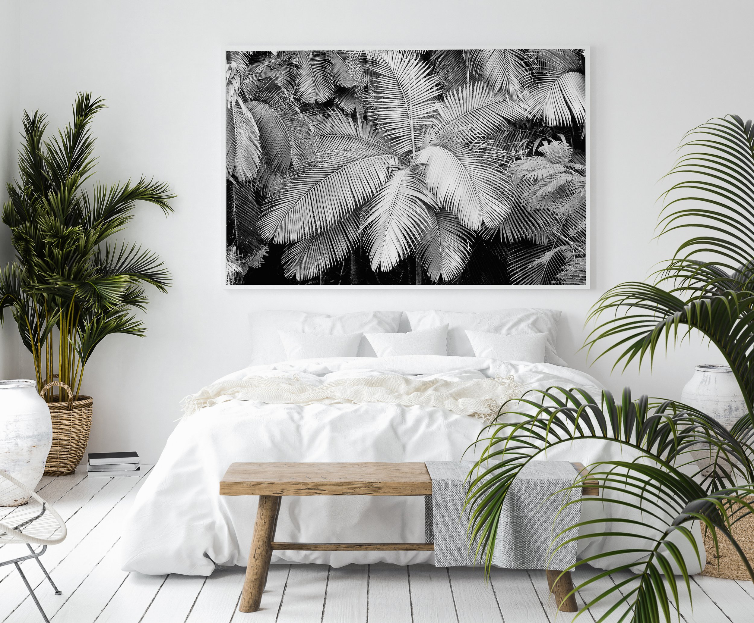 Minimalist bedroom with white walls, a large black-and-white leaf-patterned artwork, green plants, white bedsheets, and a wooden bench at the foot of the bed.