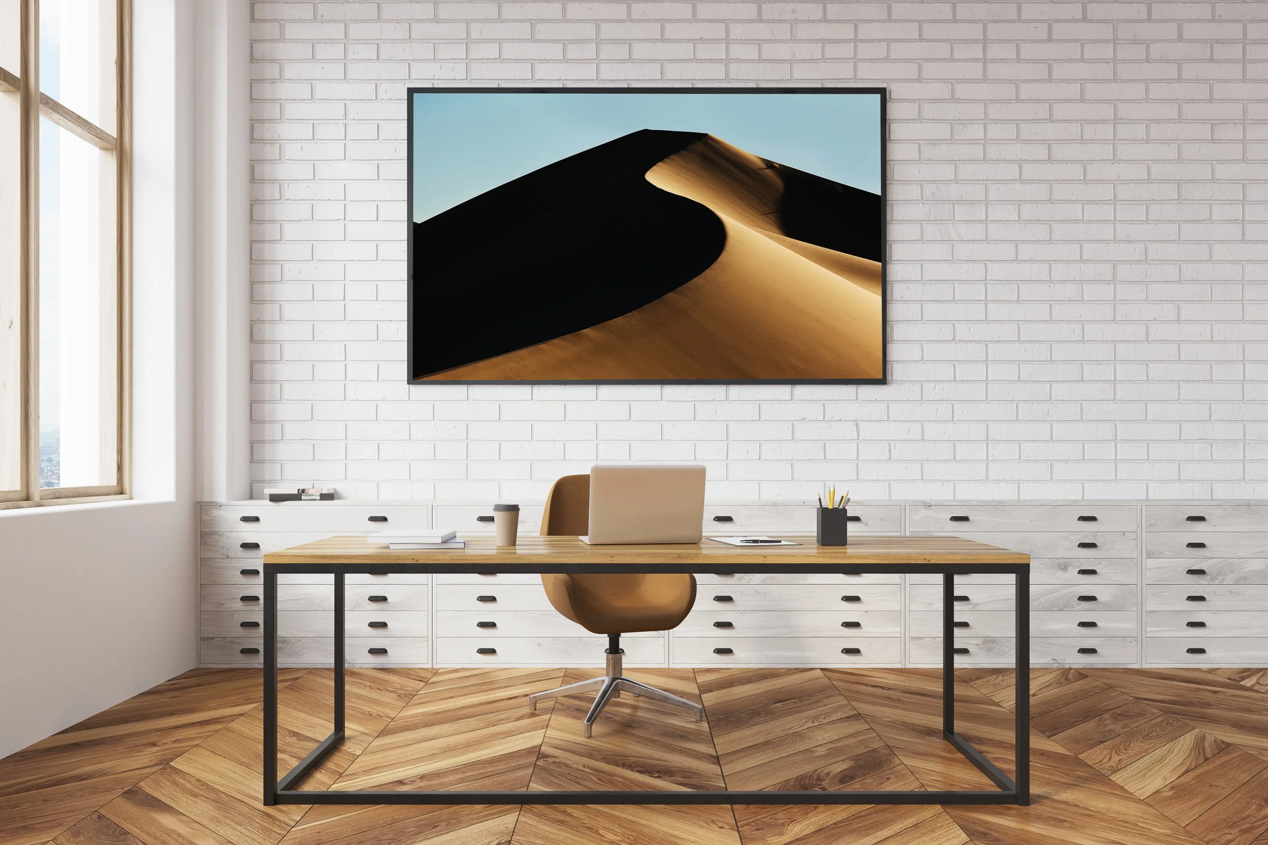 Modern office with white brick wall, large window, and wooden flooring. Desk with laptop, coffee cup, notebooks, and pen holder. Wall-mounted monitor displaying a sand dune desert landscape.
