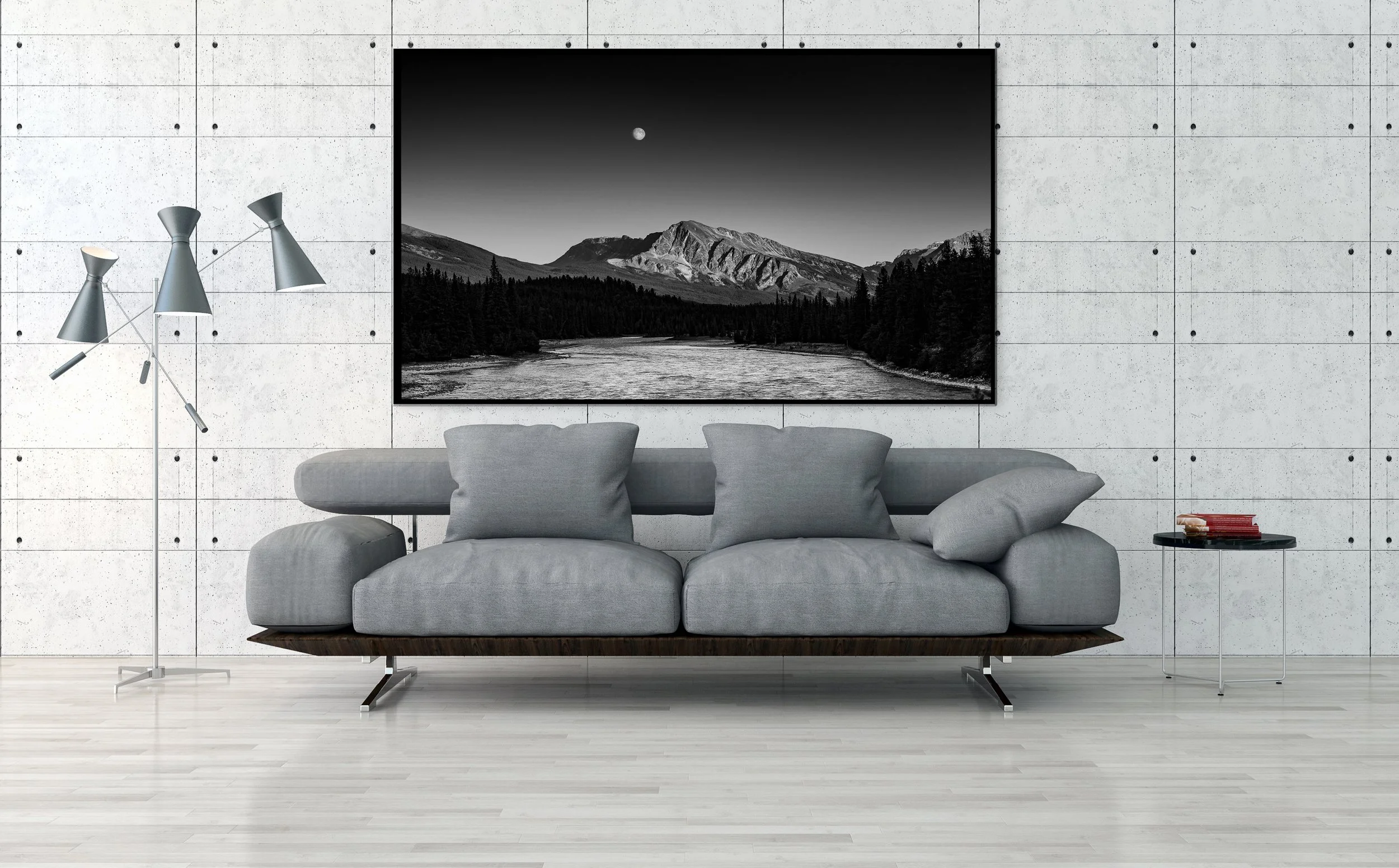 Modern living room with a gray sofa, black and white mountain landscape art on concrete wall, tripod floor lamp, and small side table with stacked books.
