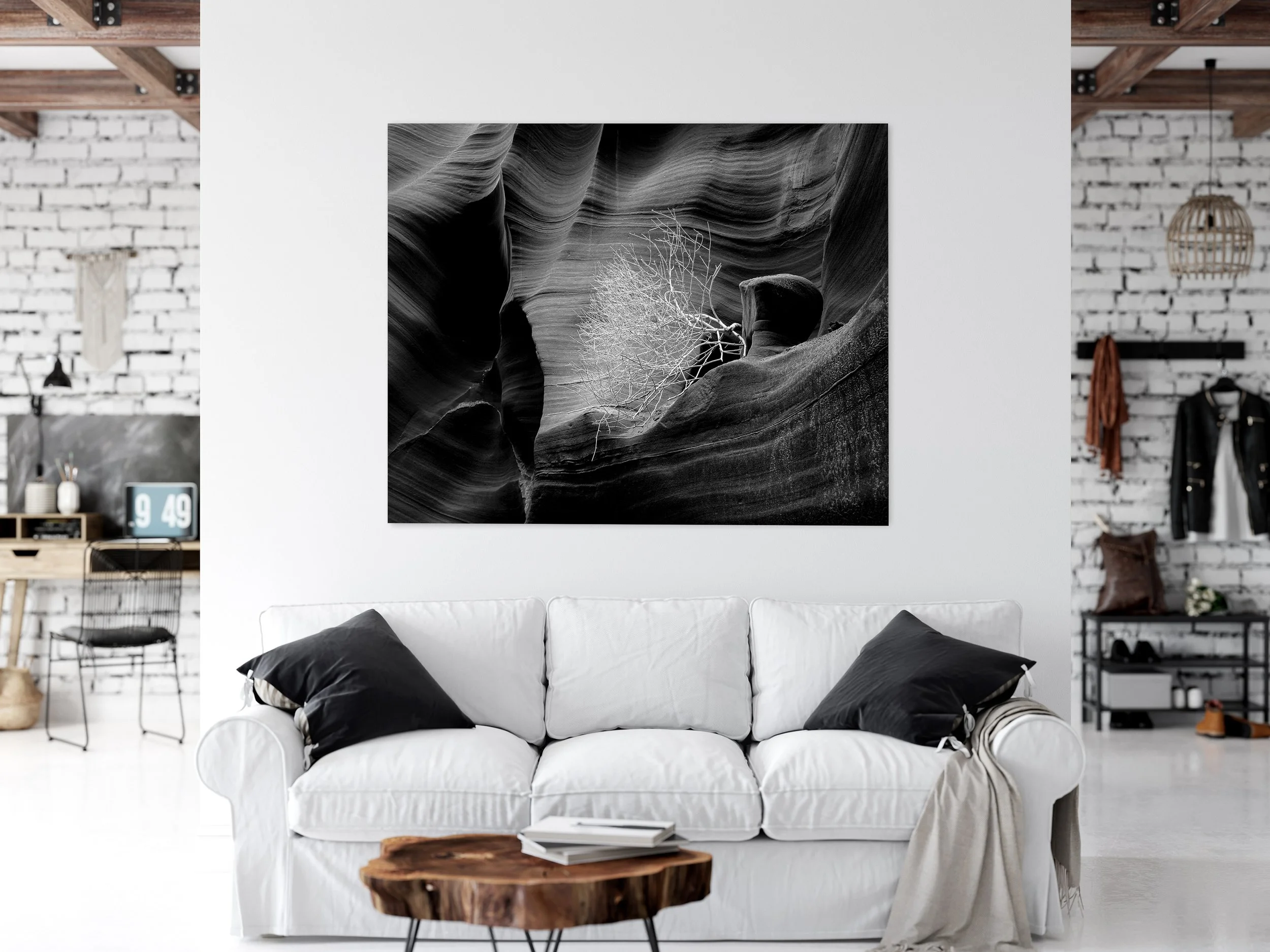 A living room with a white sofa, black pillows, large black and white photograph of a canyon hanging on the wall, wooden side table, and a background with a exposed brick wall, hanging lamps, and shelves with various decorative items.