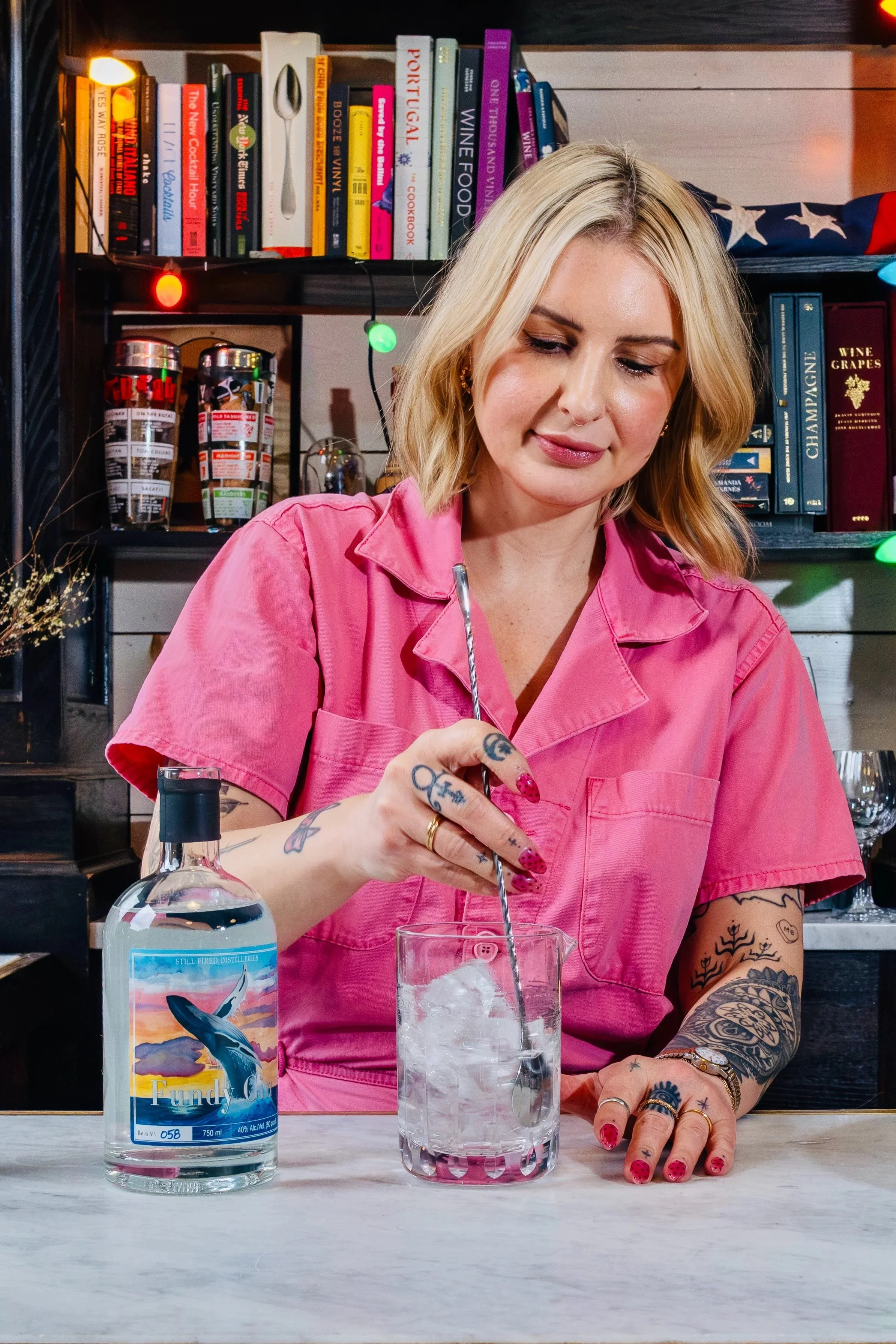 A woman with blonde hair and tattoos in a pink shirt makes a cocktail in a bar or kitchen. There is a bottle of alcohol, an ice-filled glass, and a bar with books and decorative items in the background.