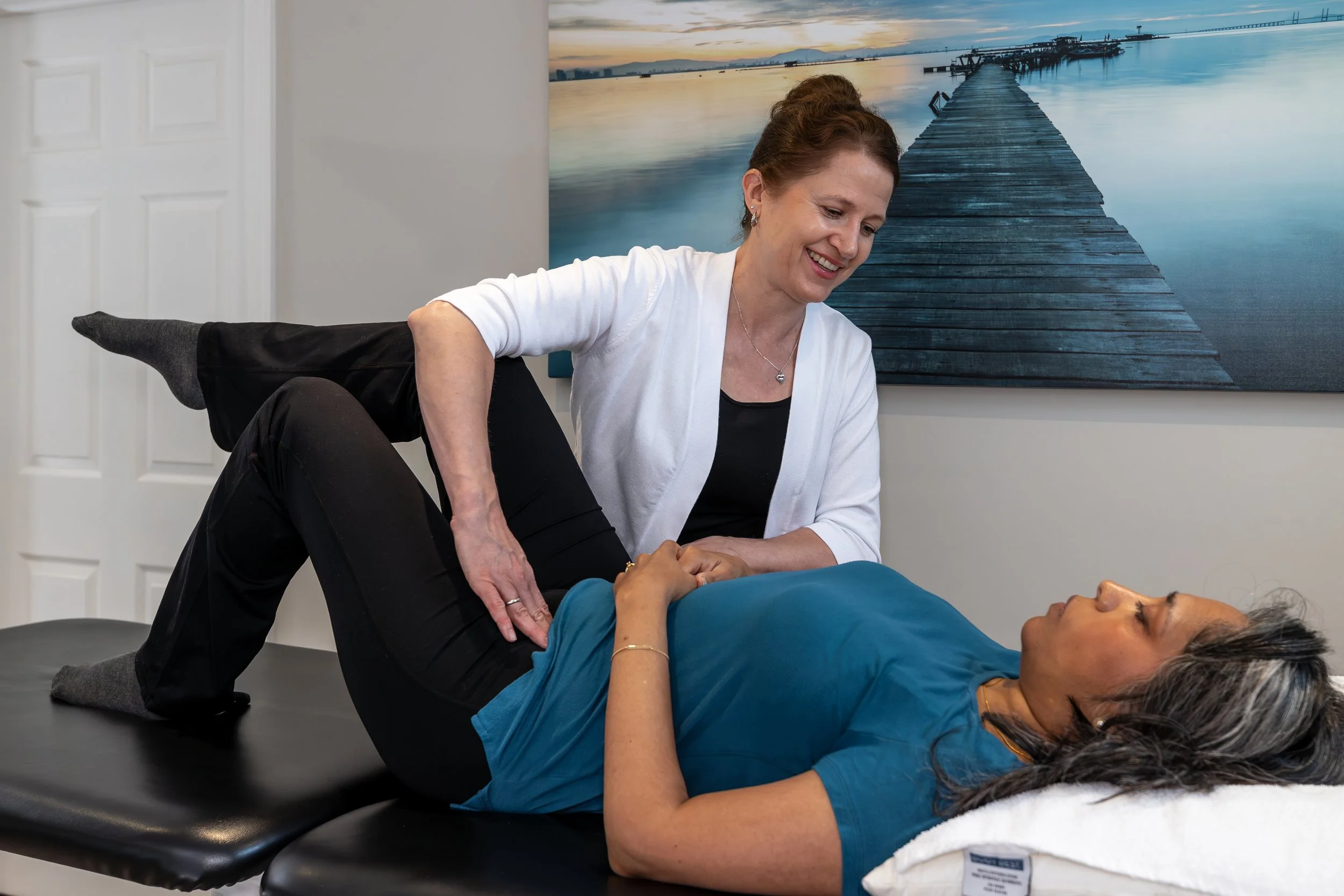 Woman performing a physical therapy exercise with another woman lying on her back on a black therapy table.