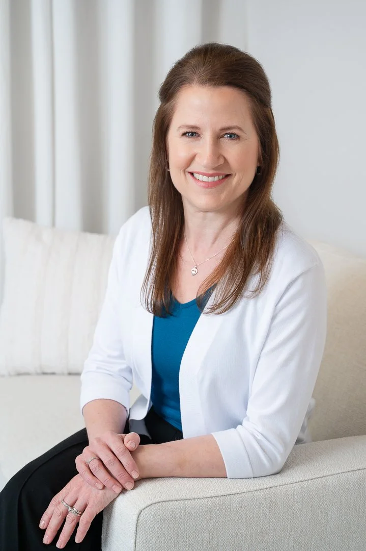 A woman with long brown hair, wearing a white blazer and blue blouse, sitting on a cream sofa, smiling in a well-lit room.