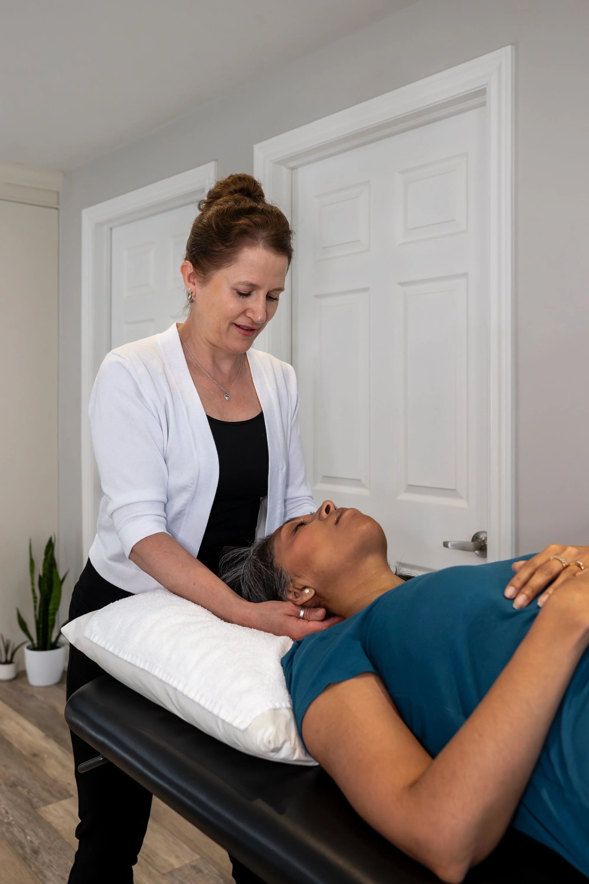 A woman is receiving a neck massage from a therapist in a treatment room with white doors and a potted plant in the background.