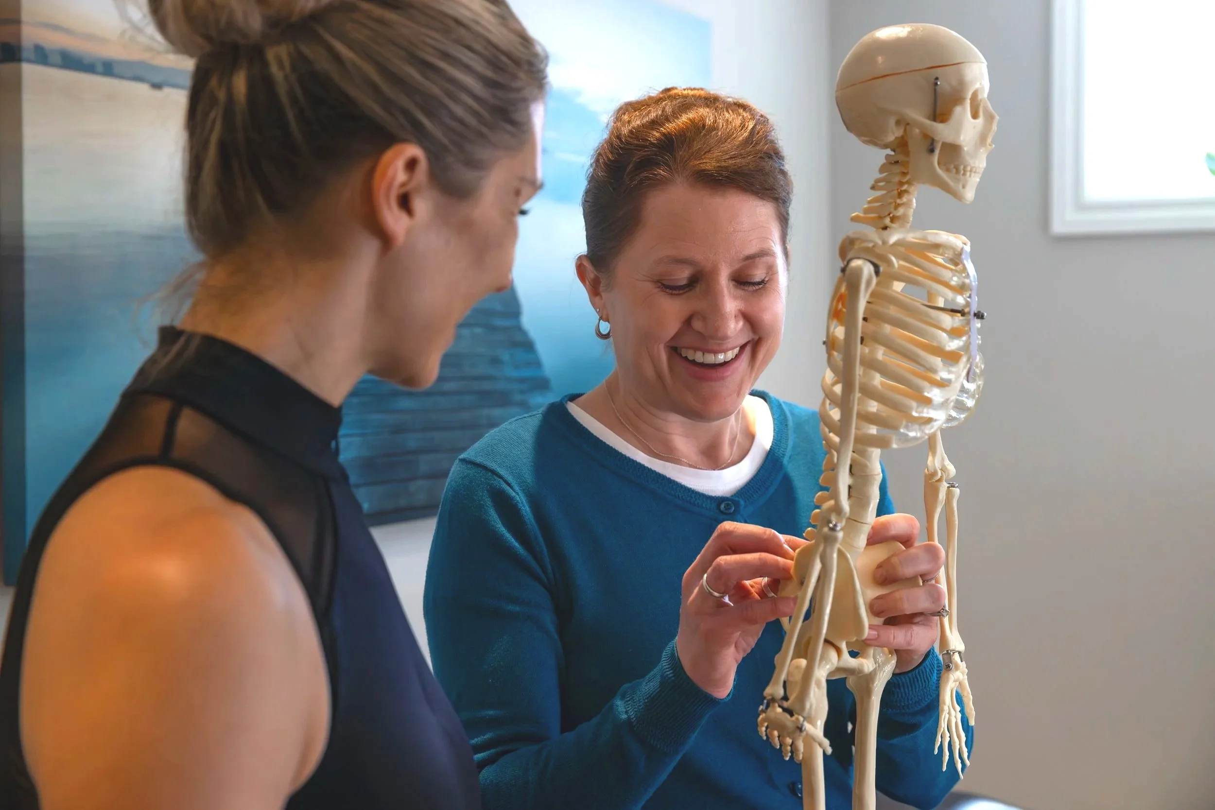 A woman is holding a human skeleton model indoors, smiling, with another woman looking on. There's a blue ocean scene in the background.