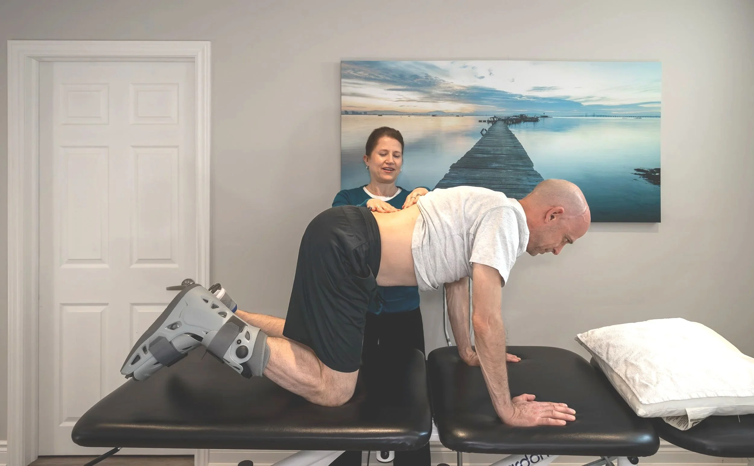 A physical therapist stabilizing a male patient in a plank position on a therapy table, with a woman assisting him, in a therapy room with a landscape painting on the wall.