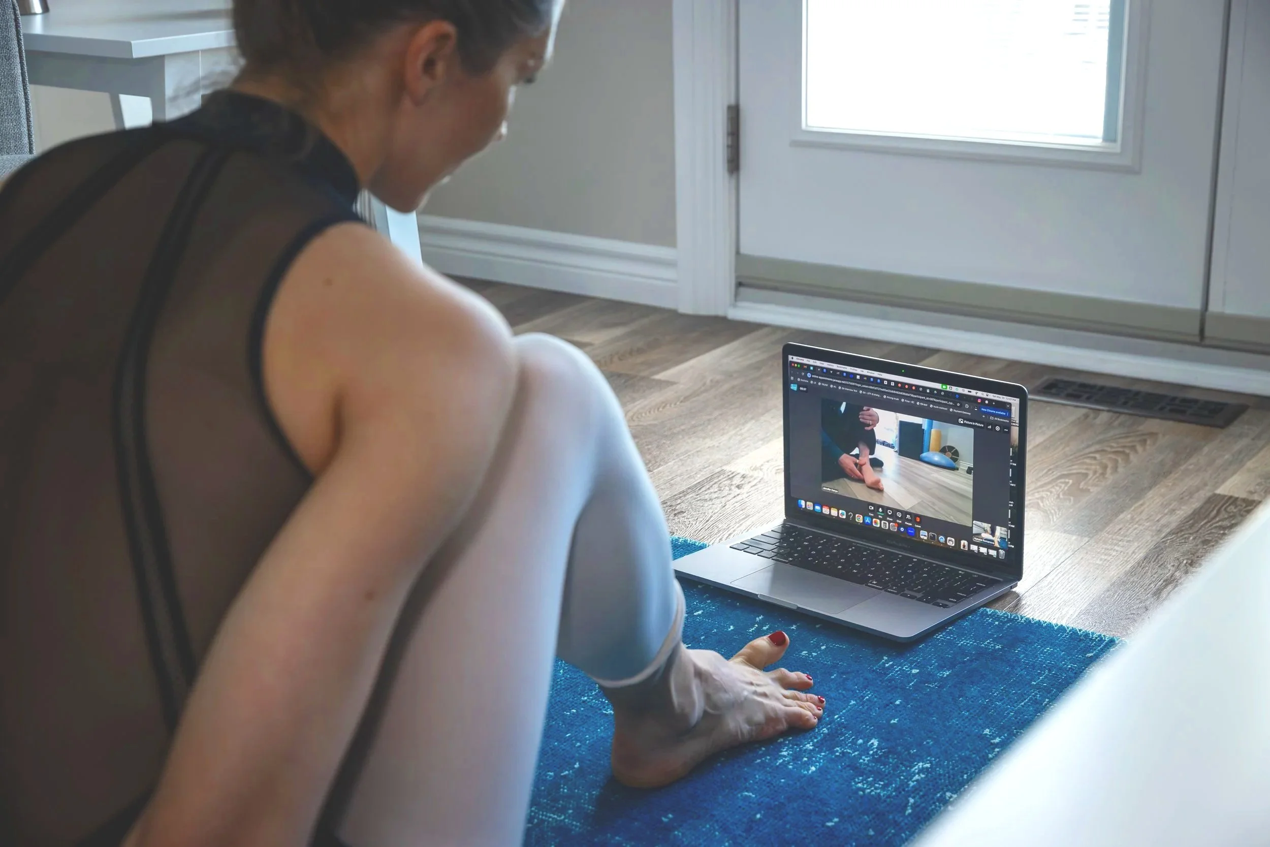 A woman in athletic wear doing a yoga pose on a blue mat inside a room, with a laptop open in front of her displaying her reflection in the screen.