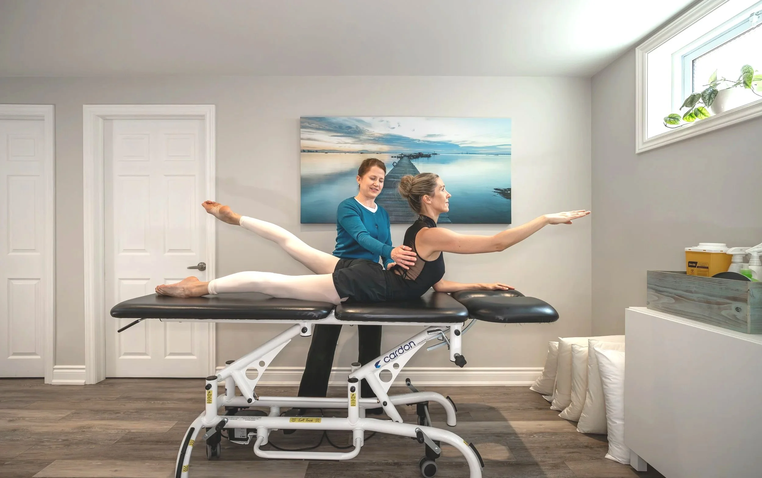 A woman is lying on her stomach on a therapy table with a pelvic correction device, while another woman, possibly a therapist, stands beside her, holding her hips, in a room with a landscape painting, a window with a plant, and medical supplies.