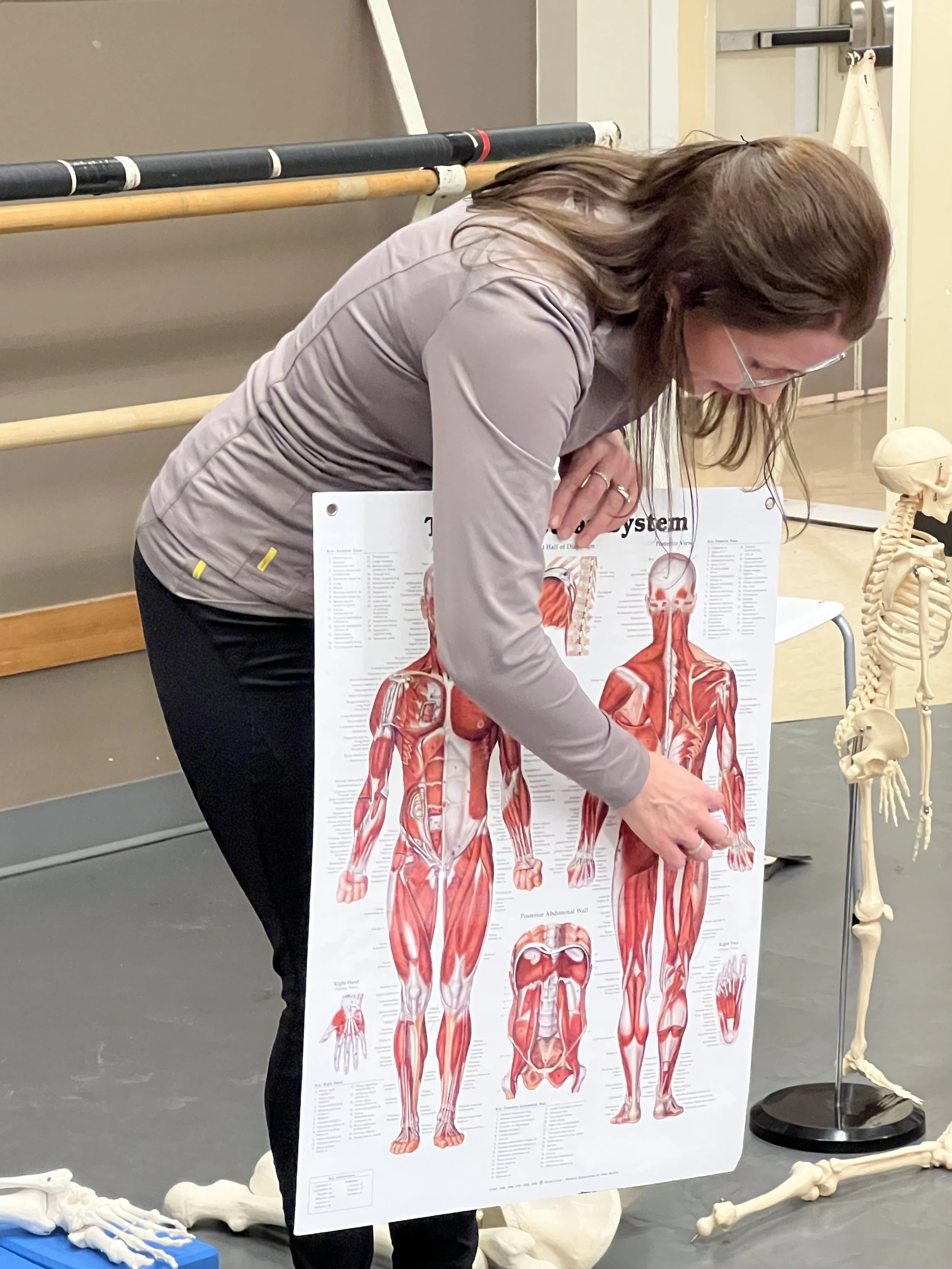 A woman with glasses pointing at a large anatomical poster of the human muscular system in a classroom or medical setting, with skeleton models on the side.