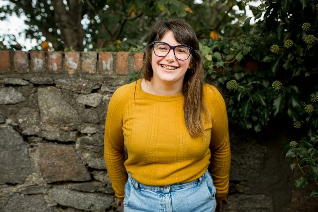 Bex is a white woman with long brown hair. She's wearing a sunny yellow top and smiling warmly.