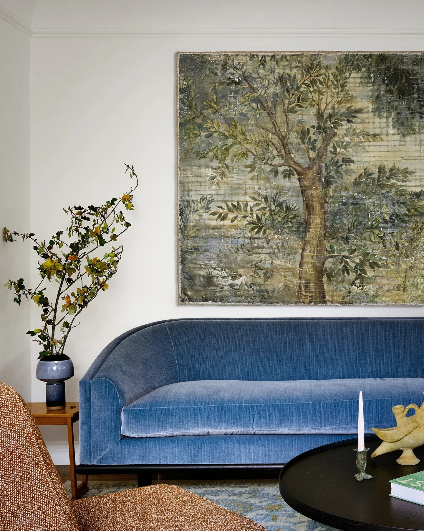 Nickey Kehoe Blue velvet sofa beneath a large painted tree artwork, with a black round coffee table and handmade ceramics