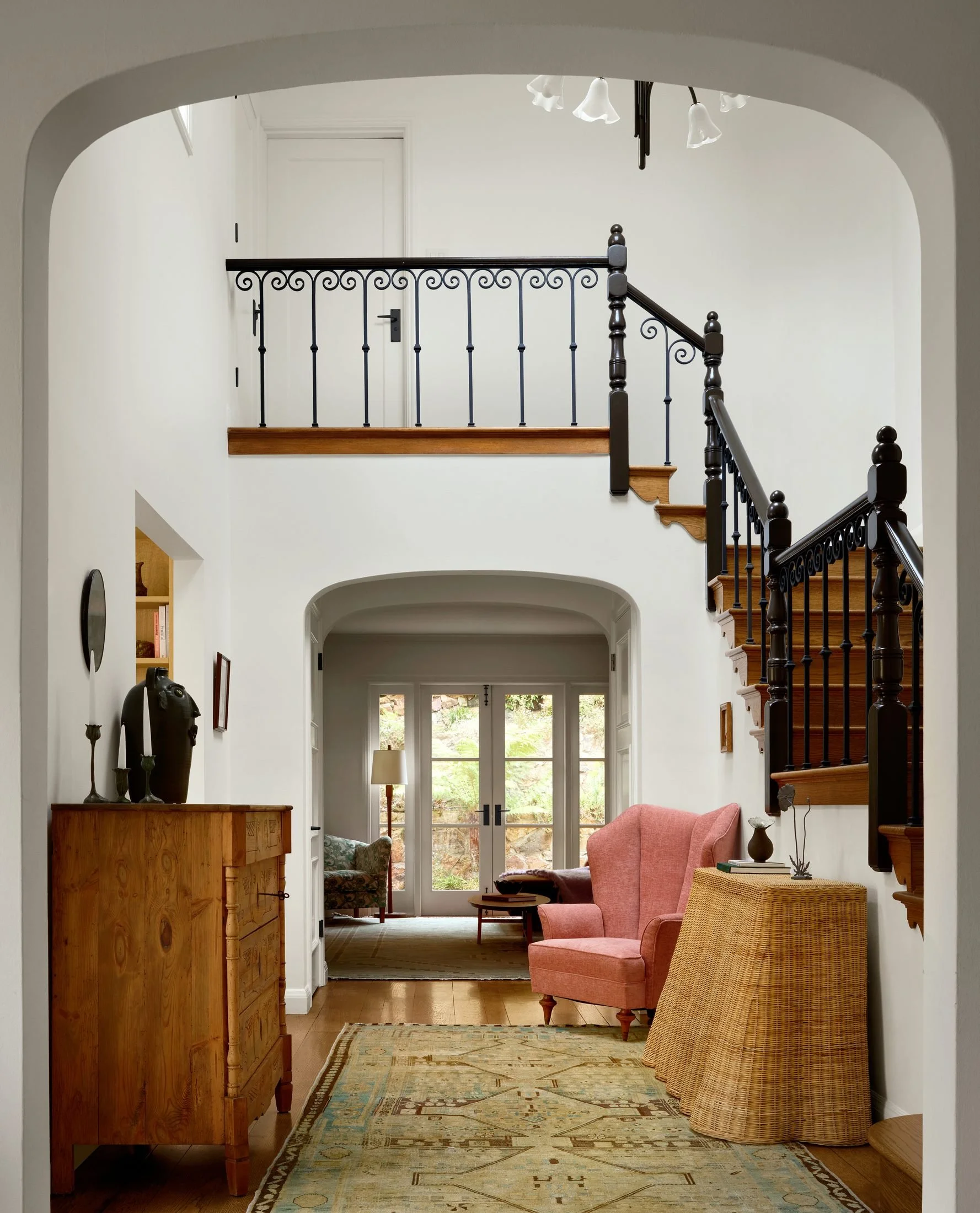 Spanish Revival foyer in San Francisco with plaster arches, wrought-iron stair, pink wingback chair and antique pine dresser