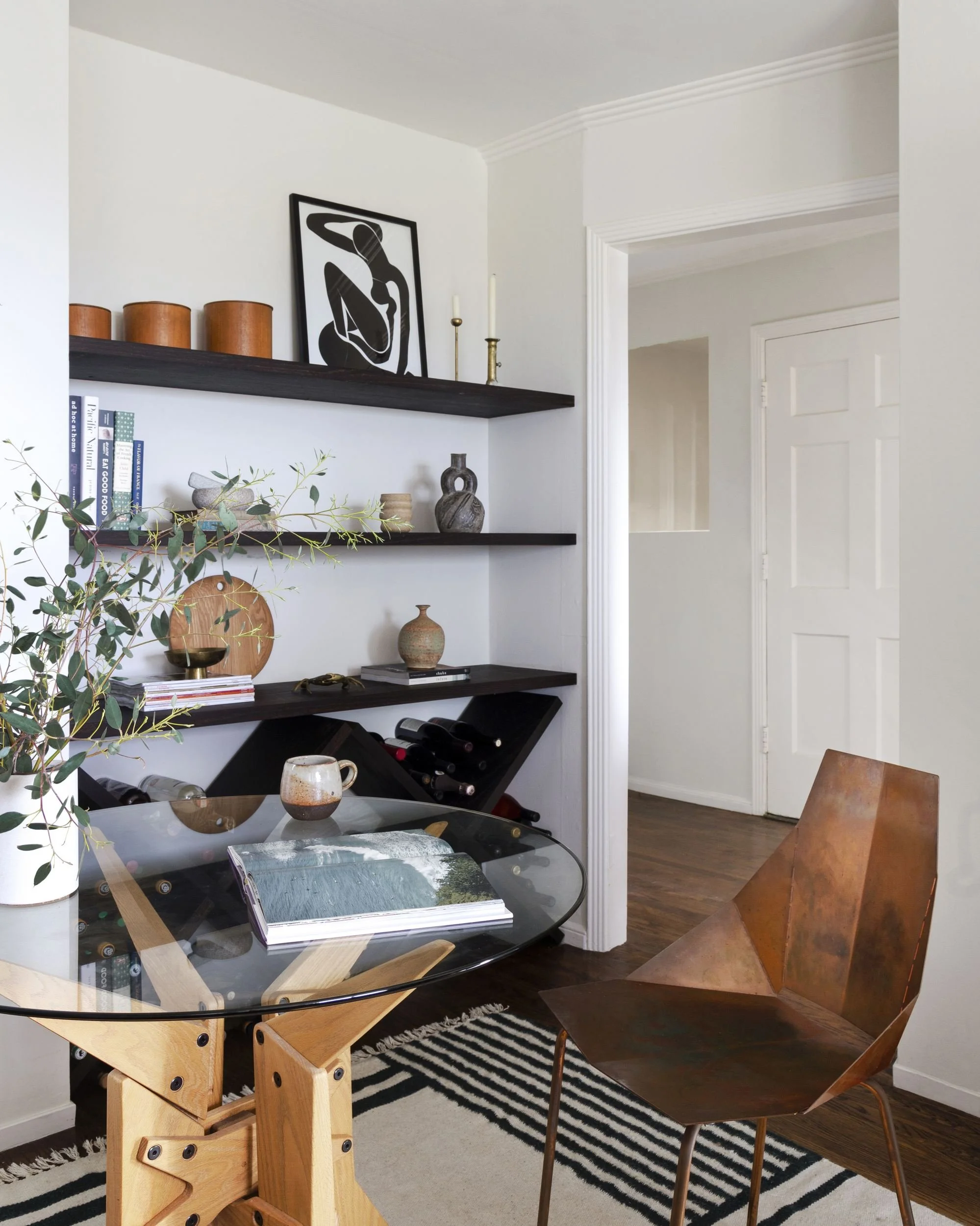 Dining corner with black open shelving, round glass table on a carved wood base and a copper faceted chair