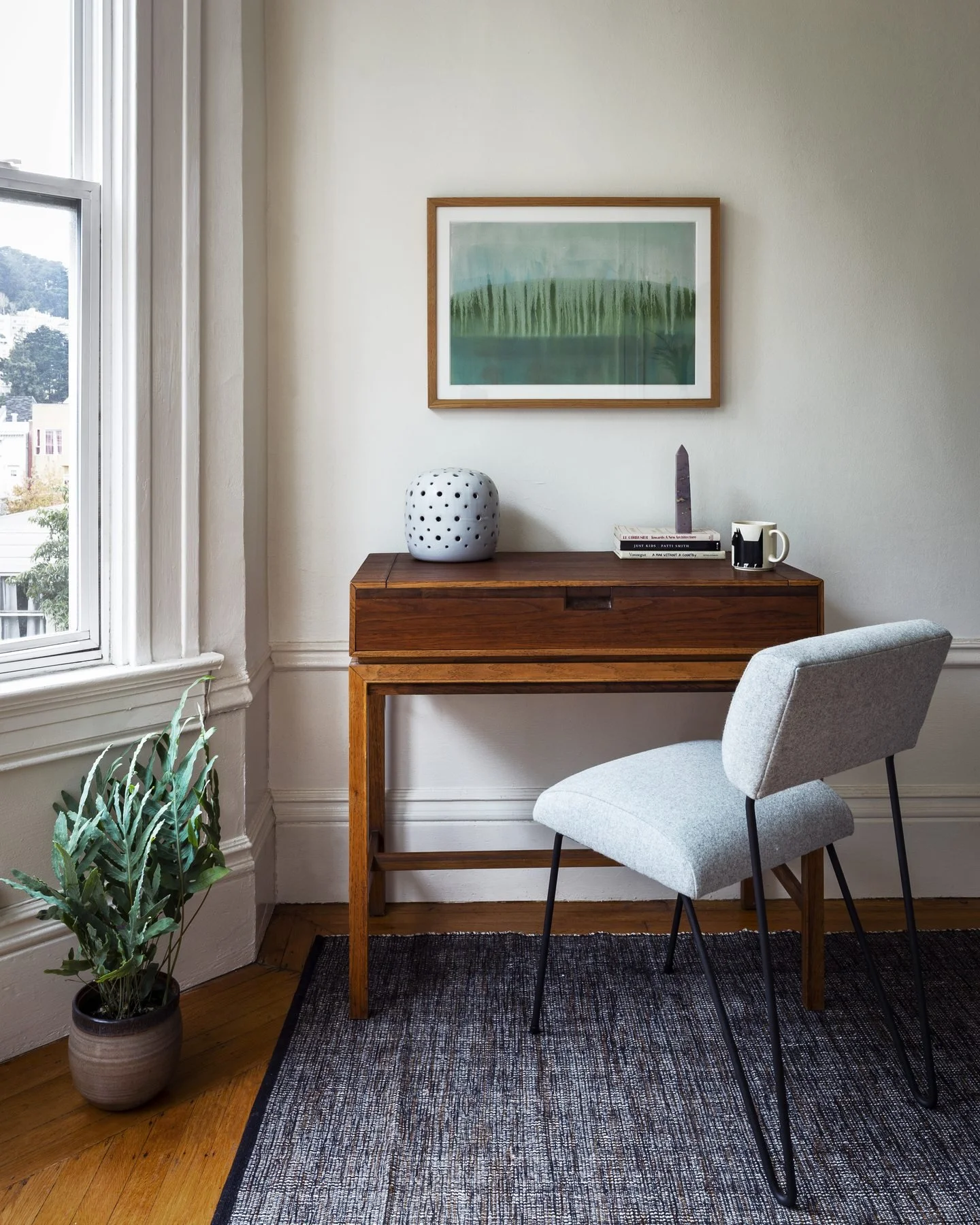 Study with a vintage wood desk, pale upholstered chair and green landscape painting by a tall sash window
