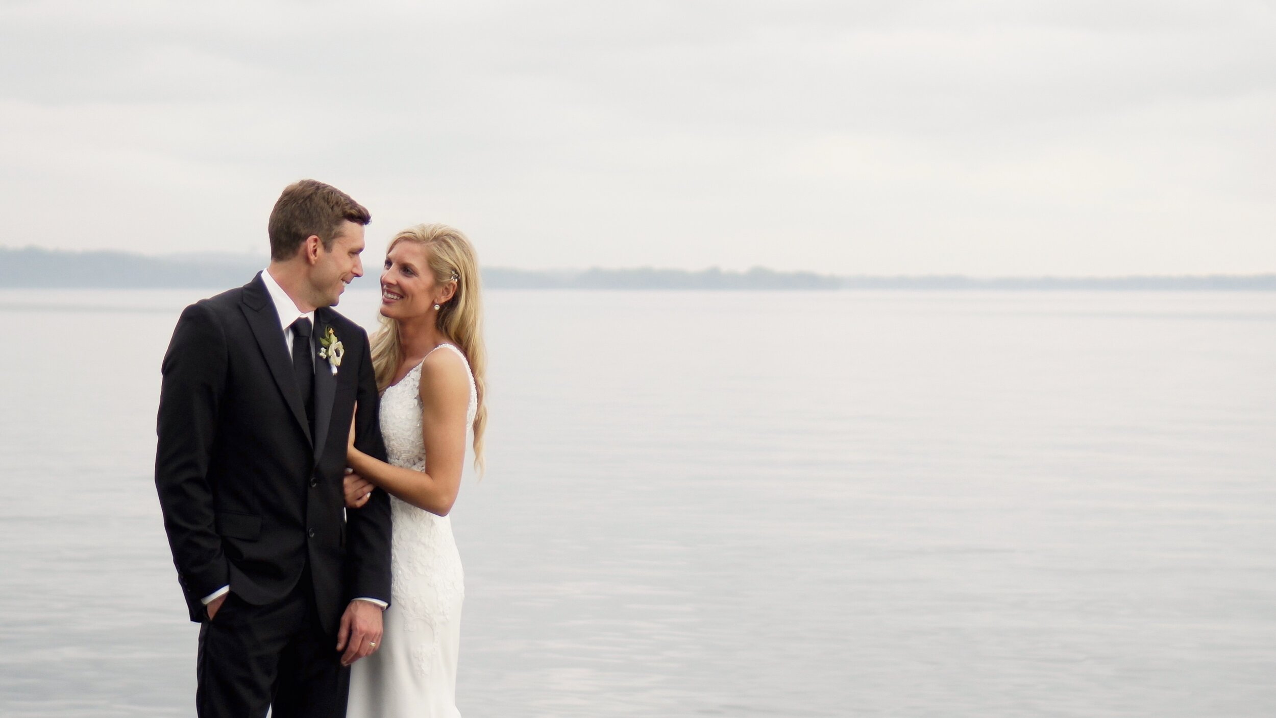 Bride in an essense of Australia dress looks at groom in black tux with a lake in the background