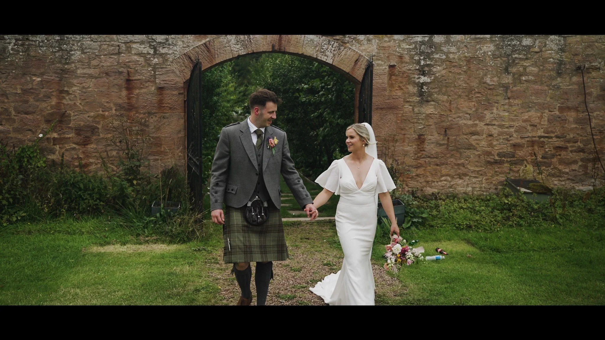 Rowan and Rhys walking hand in hand through a stone garden arch