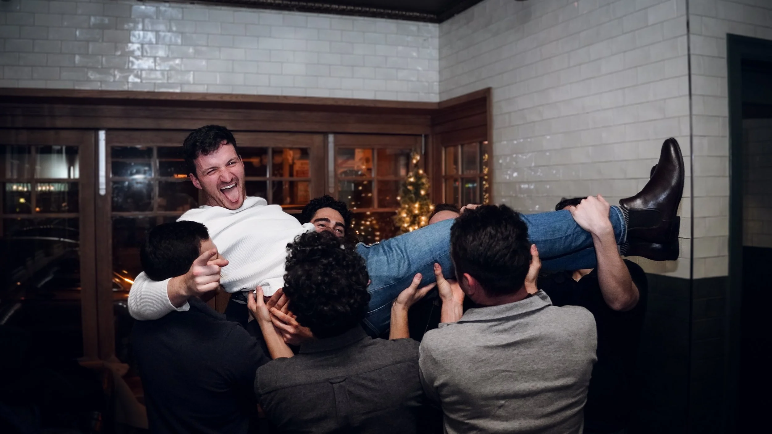 Group of friends lifting a man horizontally in a celebration setting with a Christmas tree in the background.