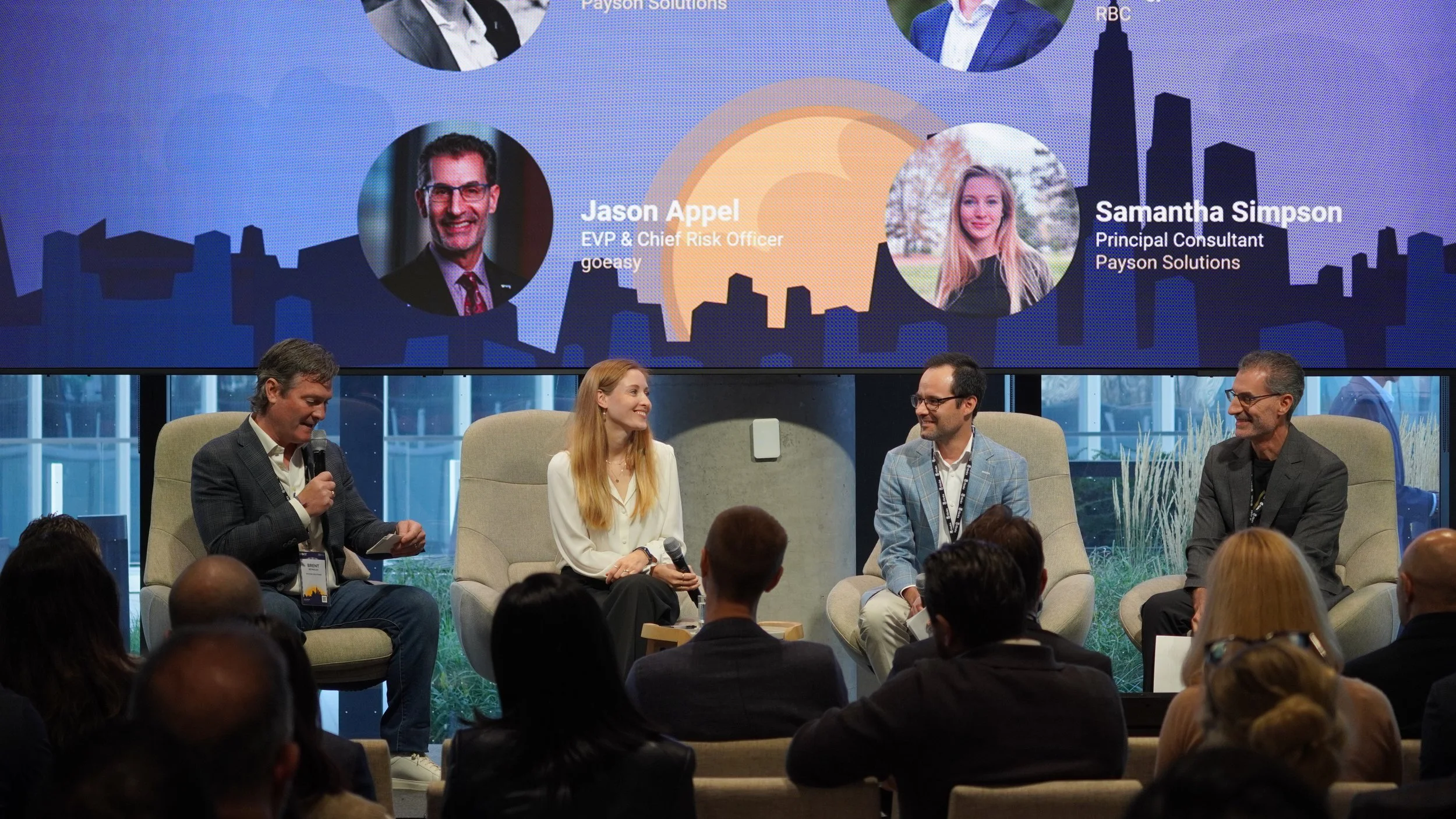 A panel discussion at a conference with four speakers seated on stage and an audience in front. Behind them, a large screen displays photos and titles of panelists, including Jason Appel and Samantha Simpson, with a city skyline background.